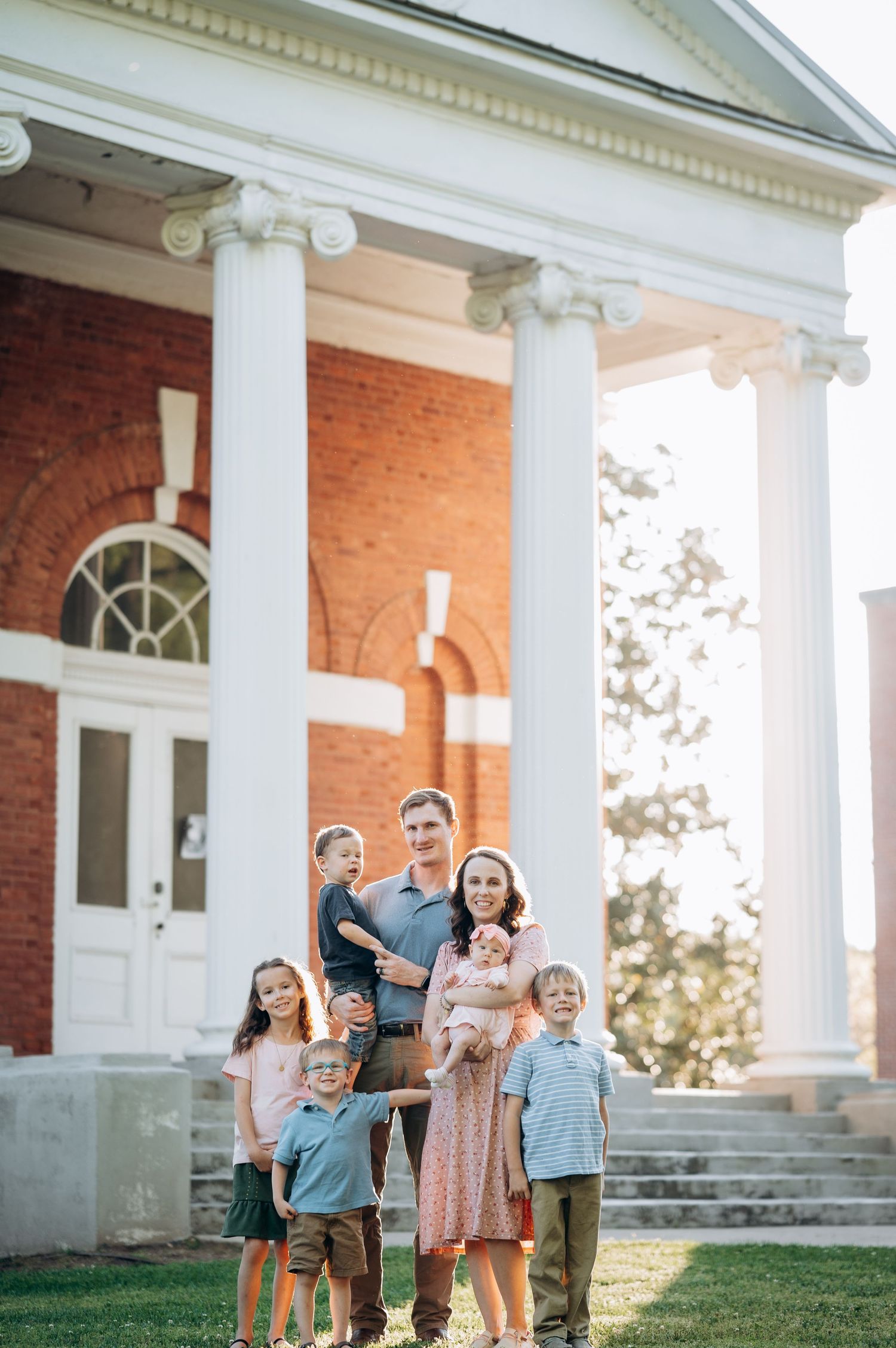 Happy family of six poses outdoors in front of a classic brick building with white columns on a sunny day.