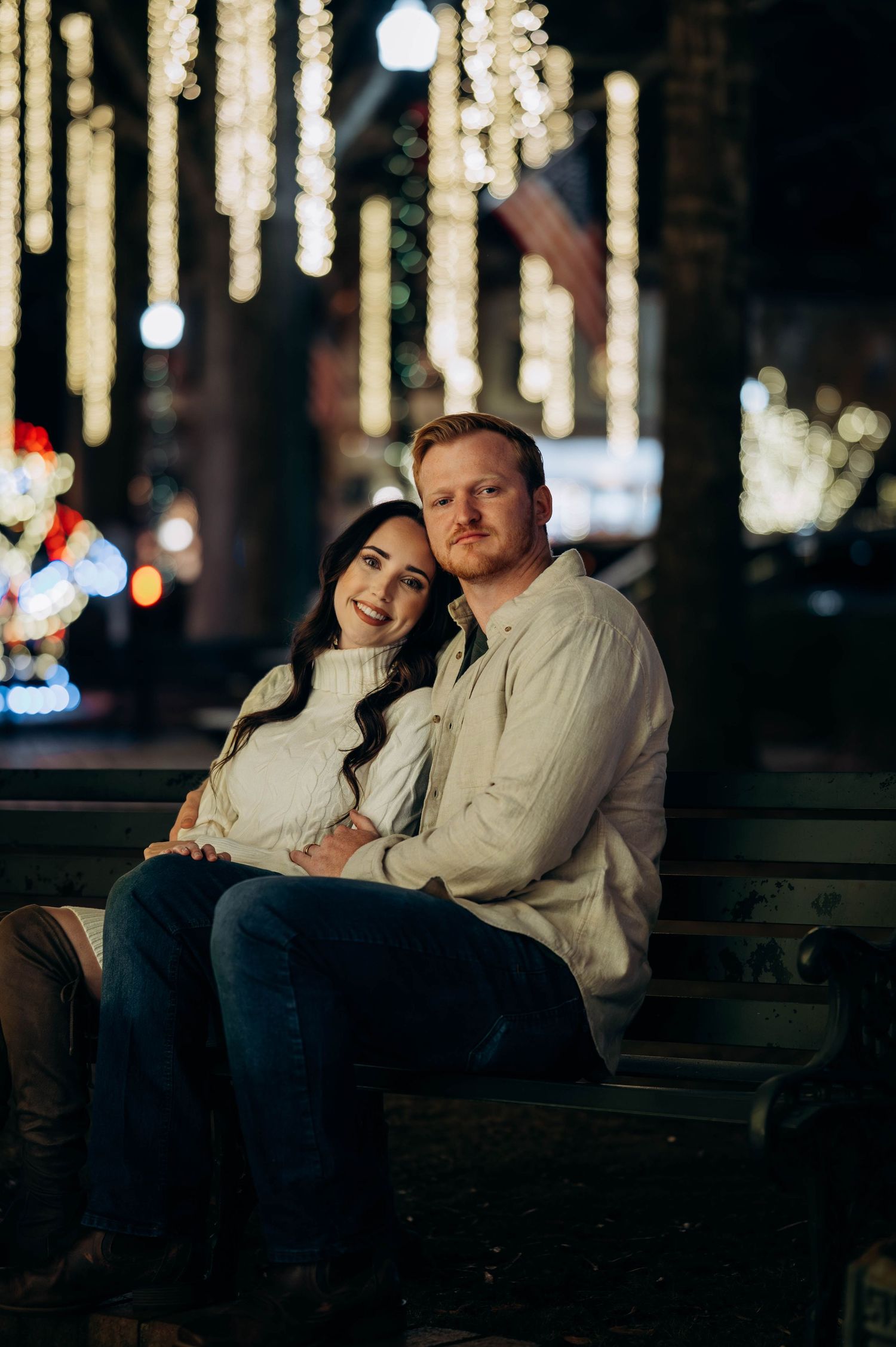 Couple sitting on a bench surrounded by glowing holiday lights at night, smiling at the camera.