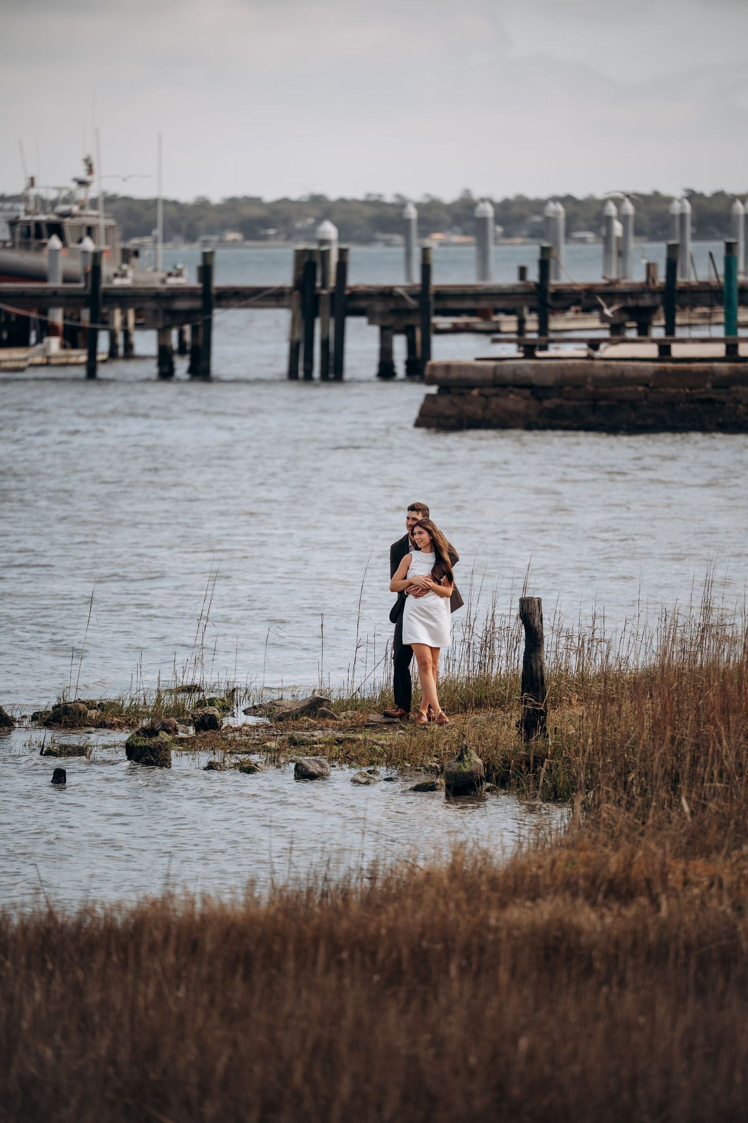 Newlyweds embrace on waterfront marsh grass near marina dock on wedding day.