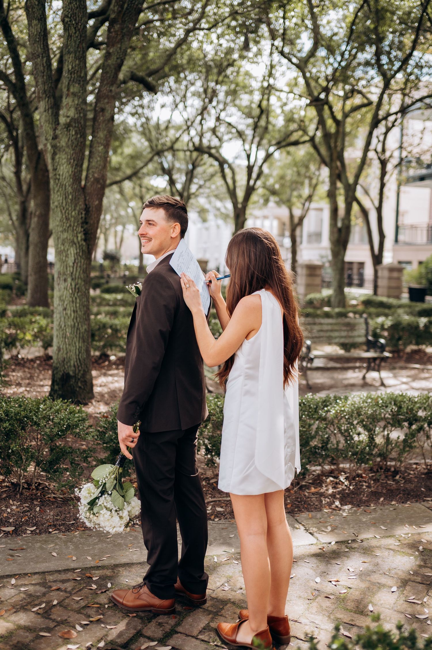 Bride adjusting groom's suit jacket in a sunlit park during a romantic wedding first look moment.