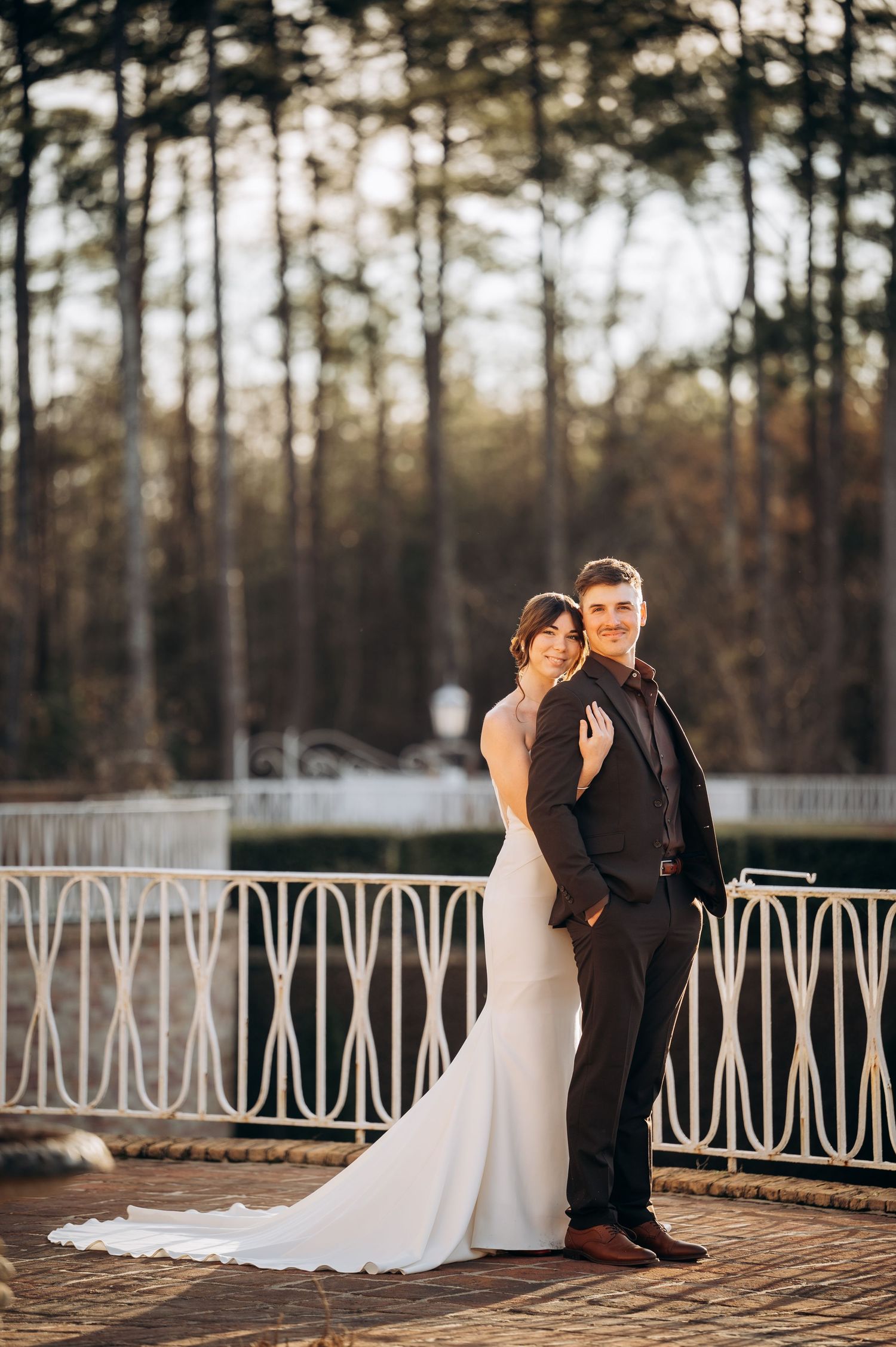 Bride and groom pose elegantly on a brick terrace with white iron railing and wooded backdrop.