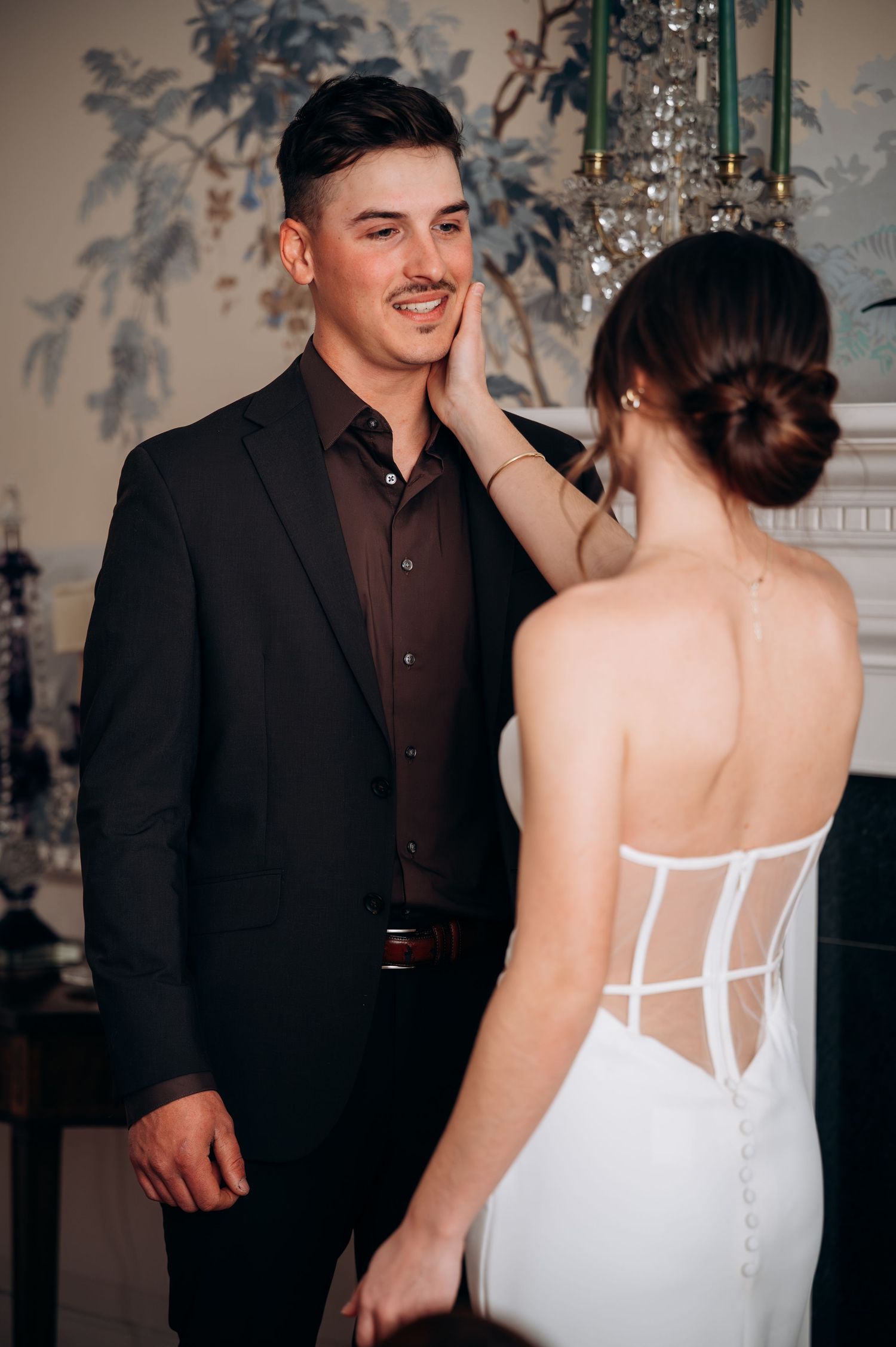 Groom in dark suit smiles as bride in corset-back white gown touches his face at wedding venue.