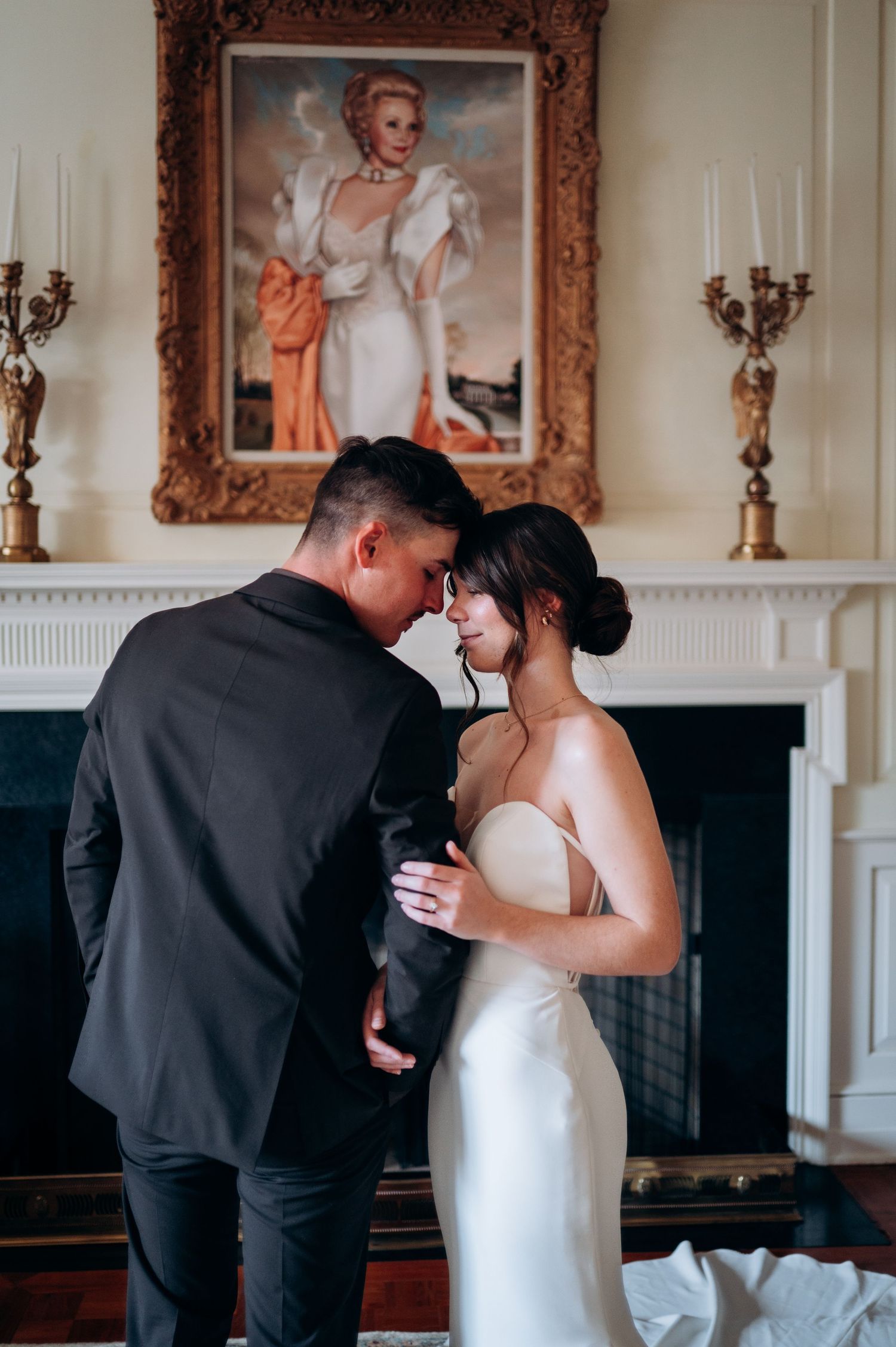Bride and groom sharing romantic forehead touch before vintage portrait painting above fireplace mantle.
