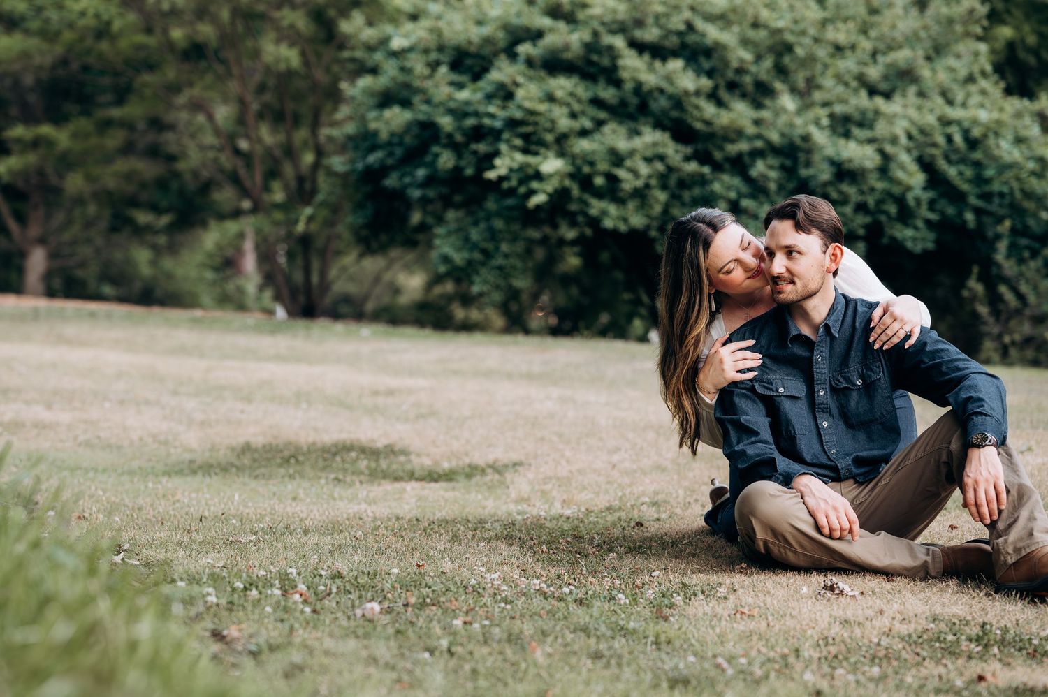 Couple sitting together on a grassy lawn, woman leaning in to kiss man on the cheek outdoors.