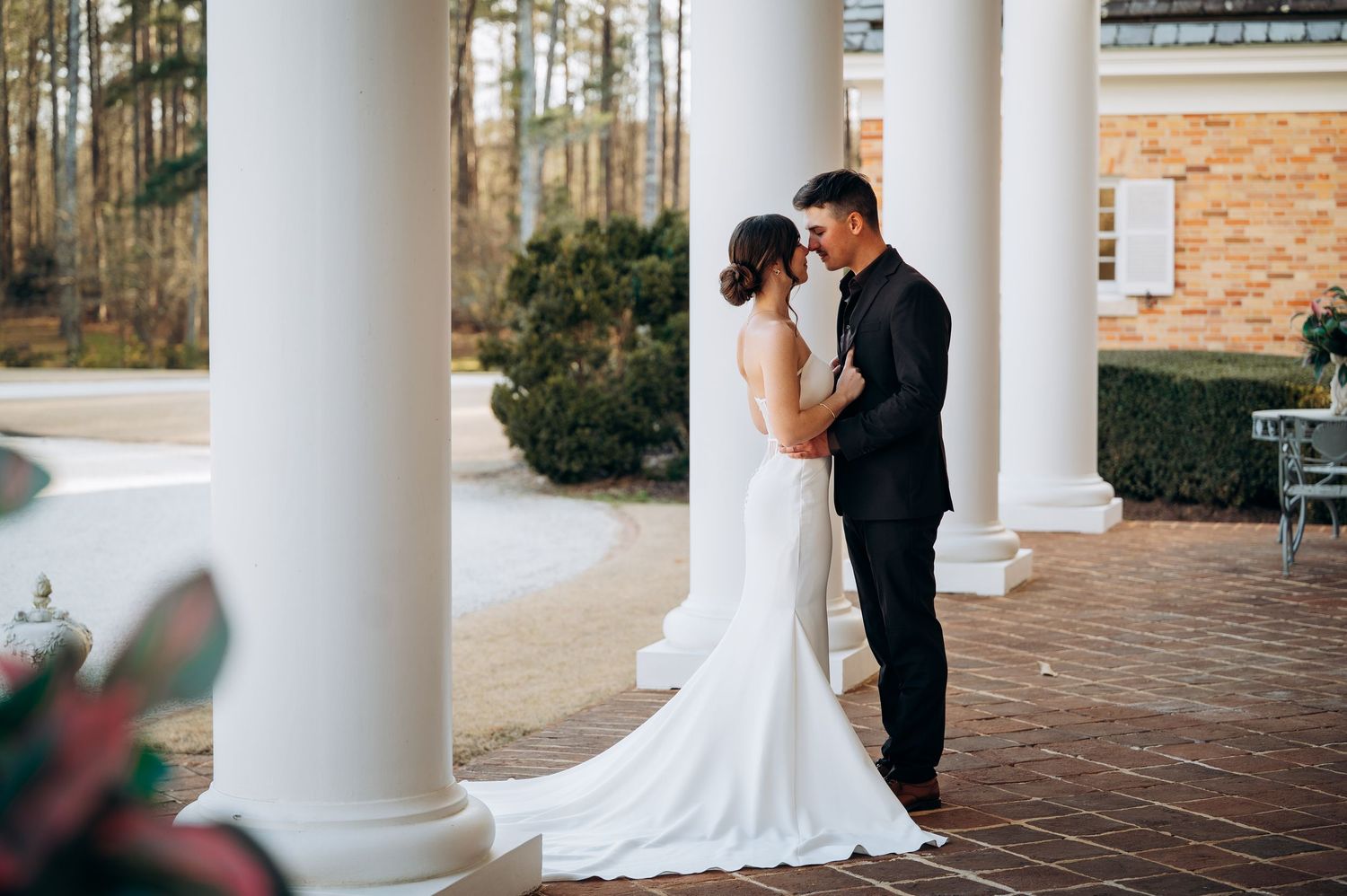 A romantic wedding moment between columns on a brick patio surrounded by greenery.