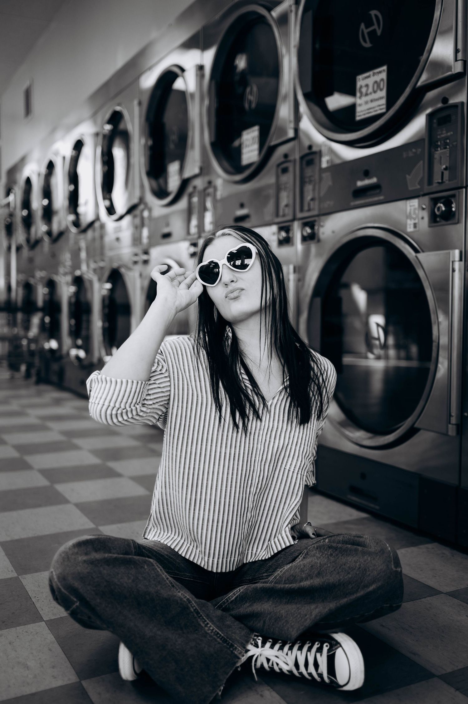 Artistic black and white photo of someone sitting cross-legged on laundromat floor wearing sunglasses.