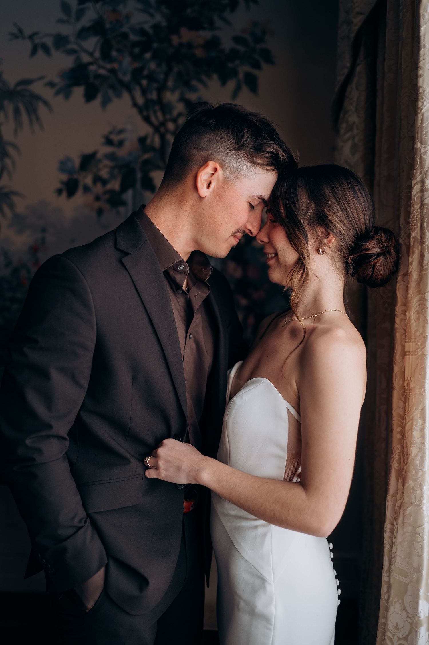 Romantic wedding couple embracing in an intimate indoor setting with natural window light.