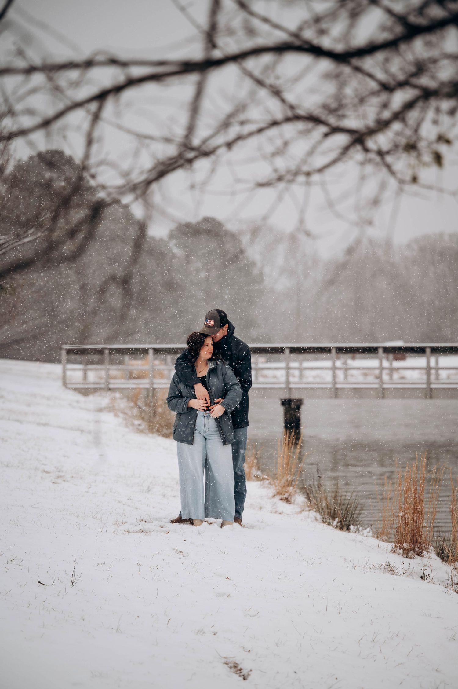 Two people share an intimate moment while standing in fresh snow with bare trees and a fence behind them.