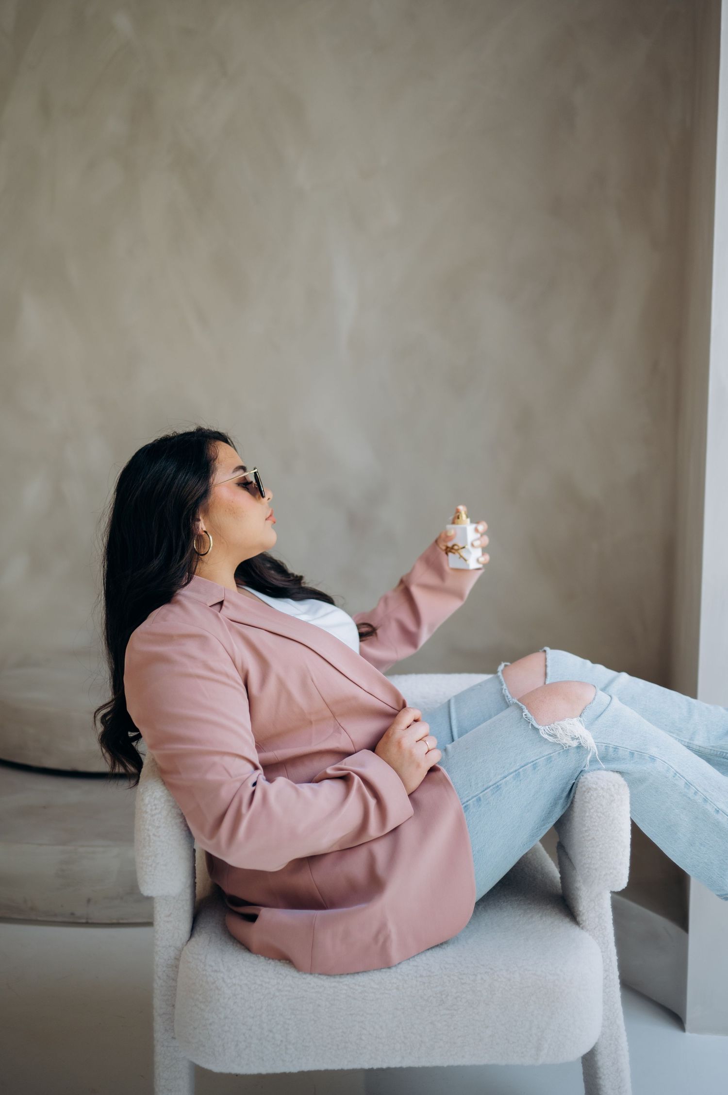 Lifestyle photo of relaxed pose in pink blazer and ripped jeans on white chair.
