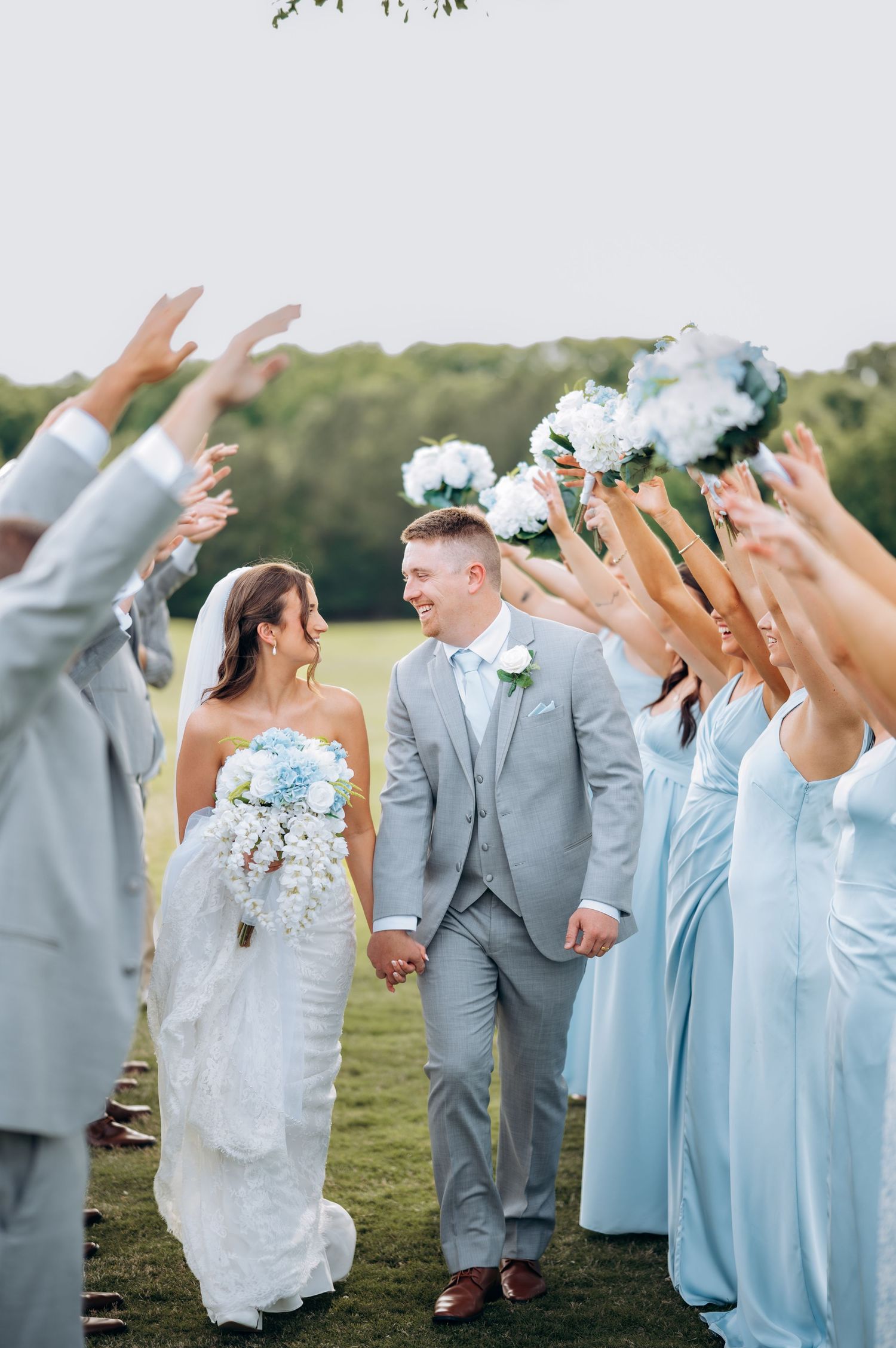 Bride and groom walk hand-in-hand through a tunnel of raised hands, surrounded by wedding party in gray and blue.