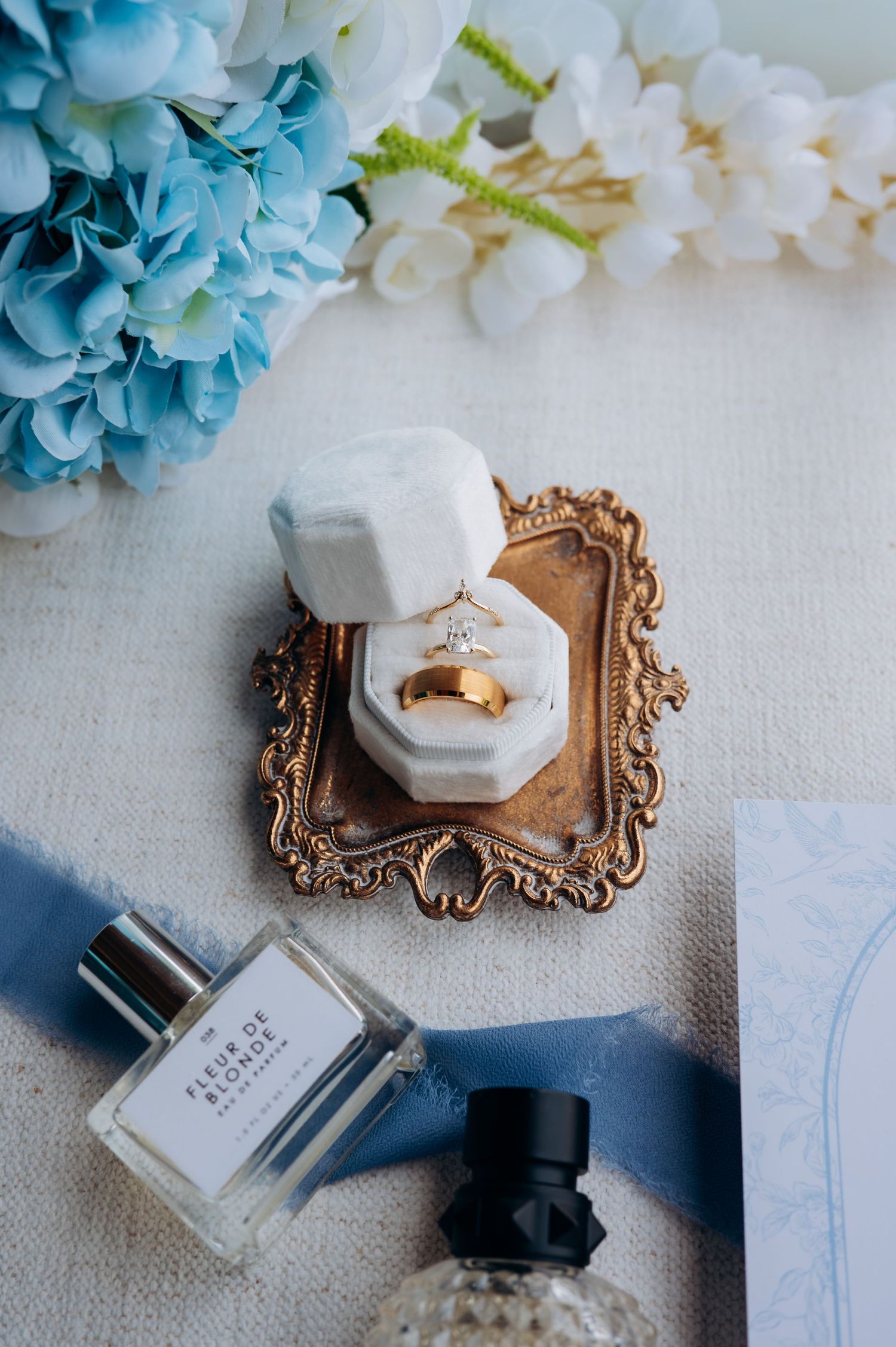 Gold wedding bands and diamond ring in white velvet box on ornate tray with blue flowers and perfume bottles.