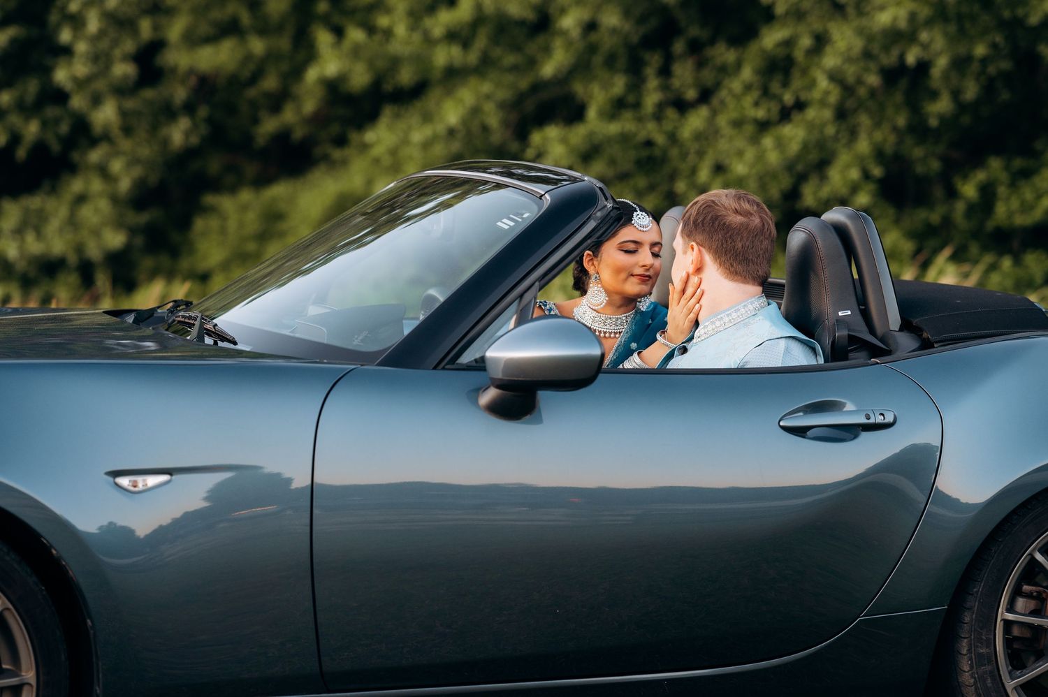 A couple sharing a romantic moment in a sleek gray convertible sports car surrounded by lush green trees.