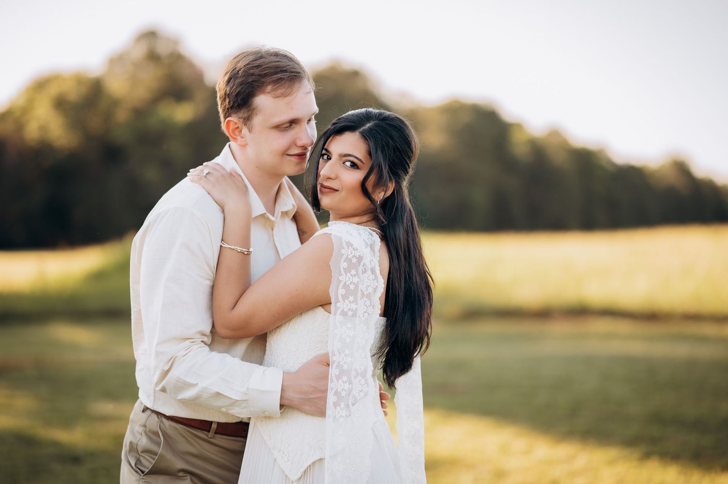 A couple embraces in a sunlit field, the bride in white lace and groom in casual attire, sharing a romantic outdoor moment.