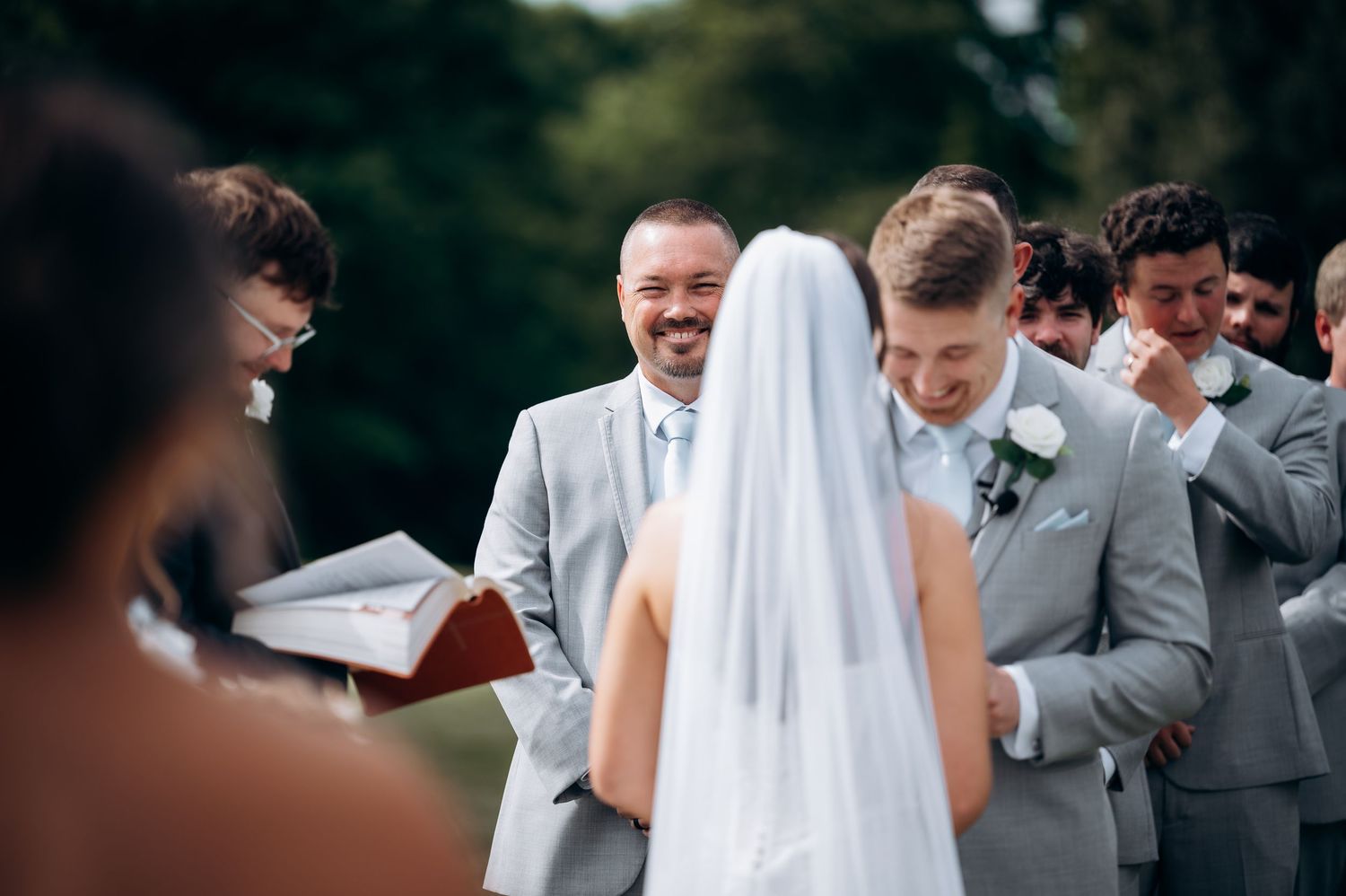 Emotional groom smiles as bride approaches the altar during outdoor wedding ceremony with groomsmen.