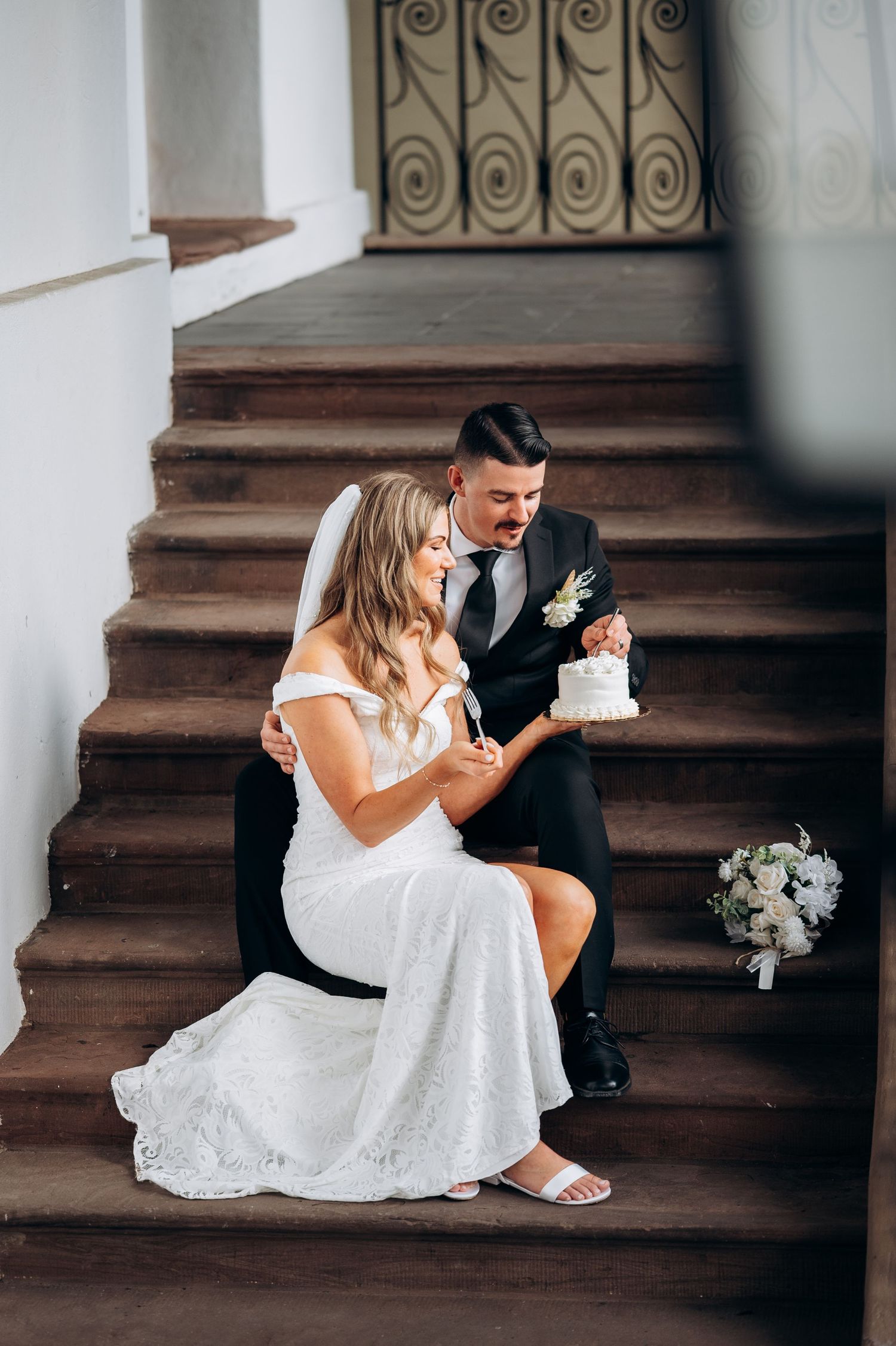 Bride and groom sitting on wooden stairs sharing a small white wedding cake.