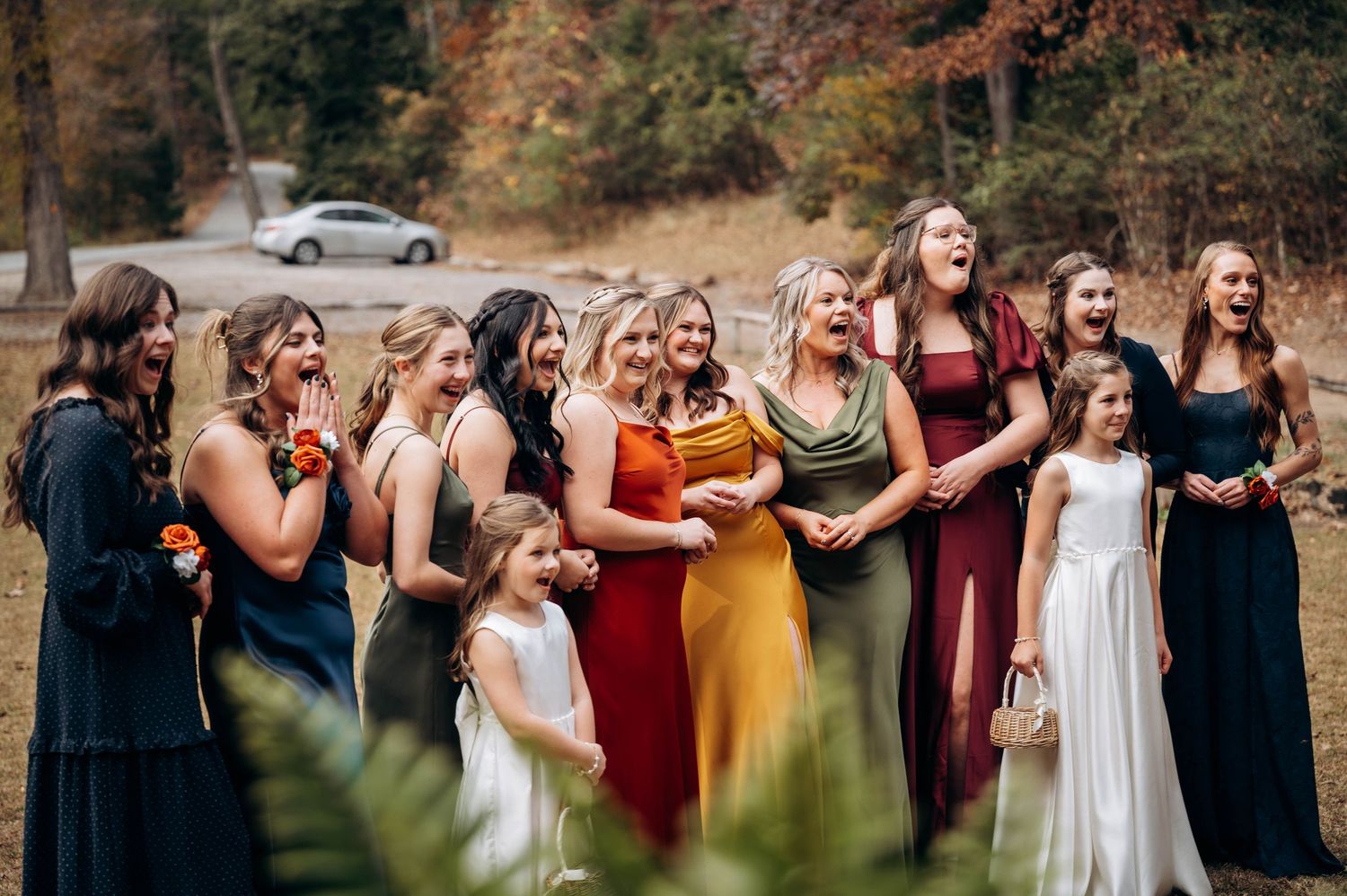 Wedding party lined up outdoors wearing mix of burgundy, white, and navy dresses against fall foliage.