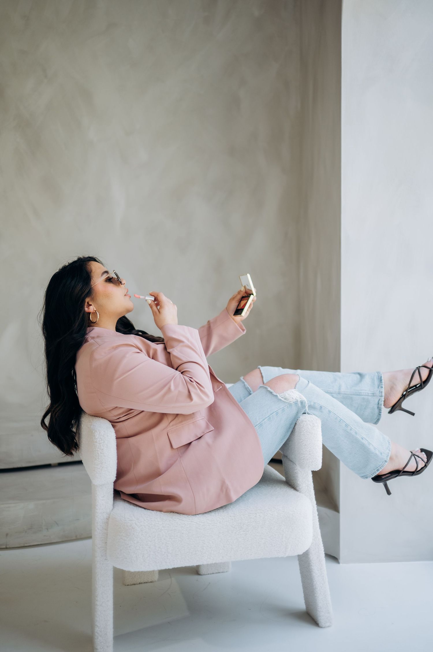 Artistic lifestyle photo showing relaxed pose applying makeup in pink blazer on white chair.