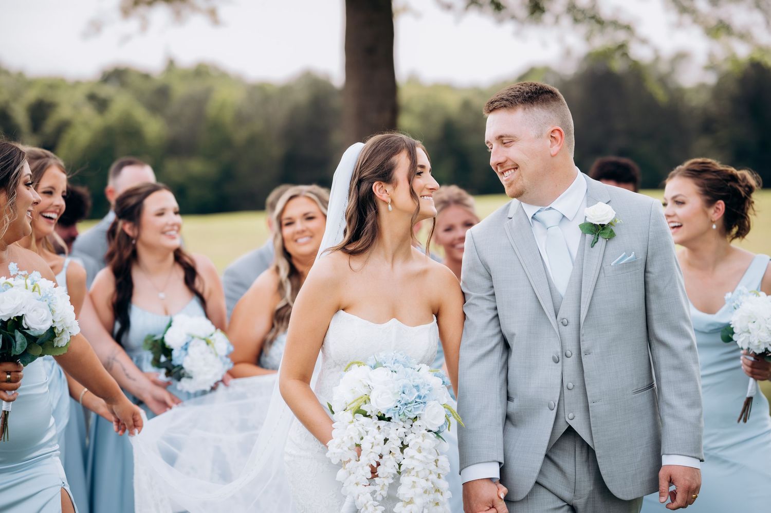 Bride and groom hold hands smiling with bridesmaids in light blue dresses at outdoor wedding celebration.