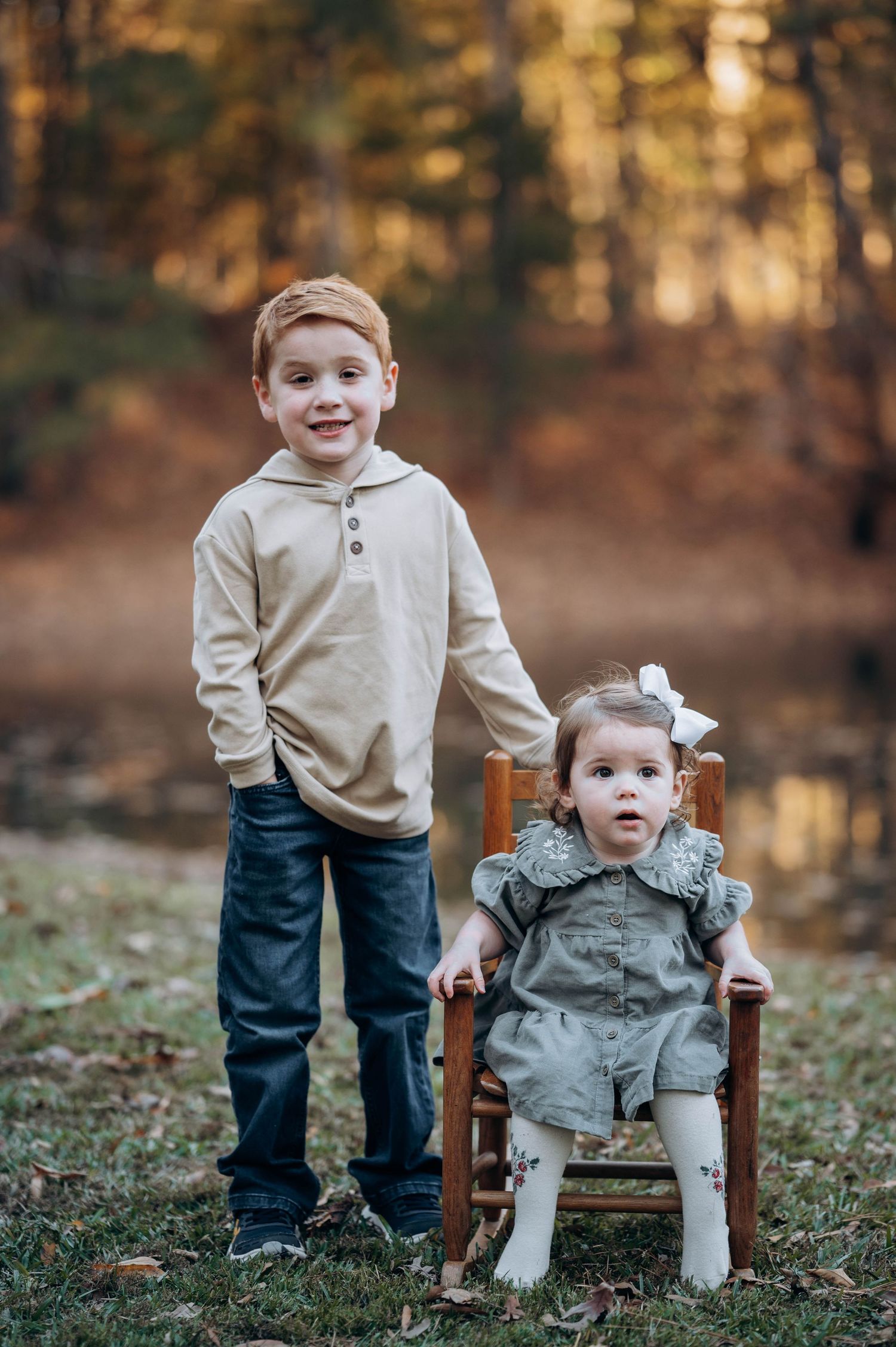 Young child sits in a wooden chair outdoors during autumn wearing cream sweater and dark pants.
