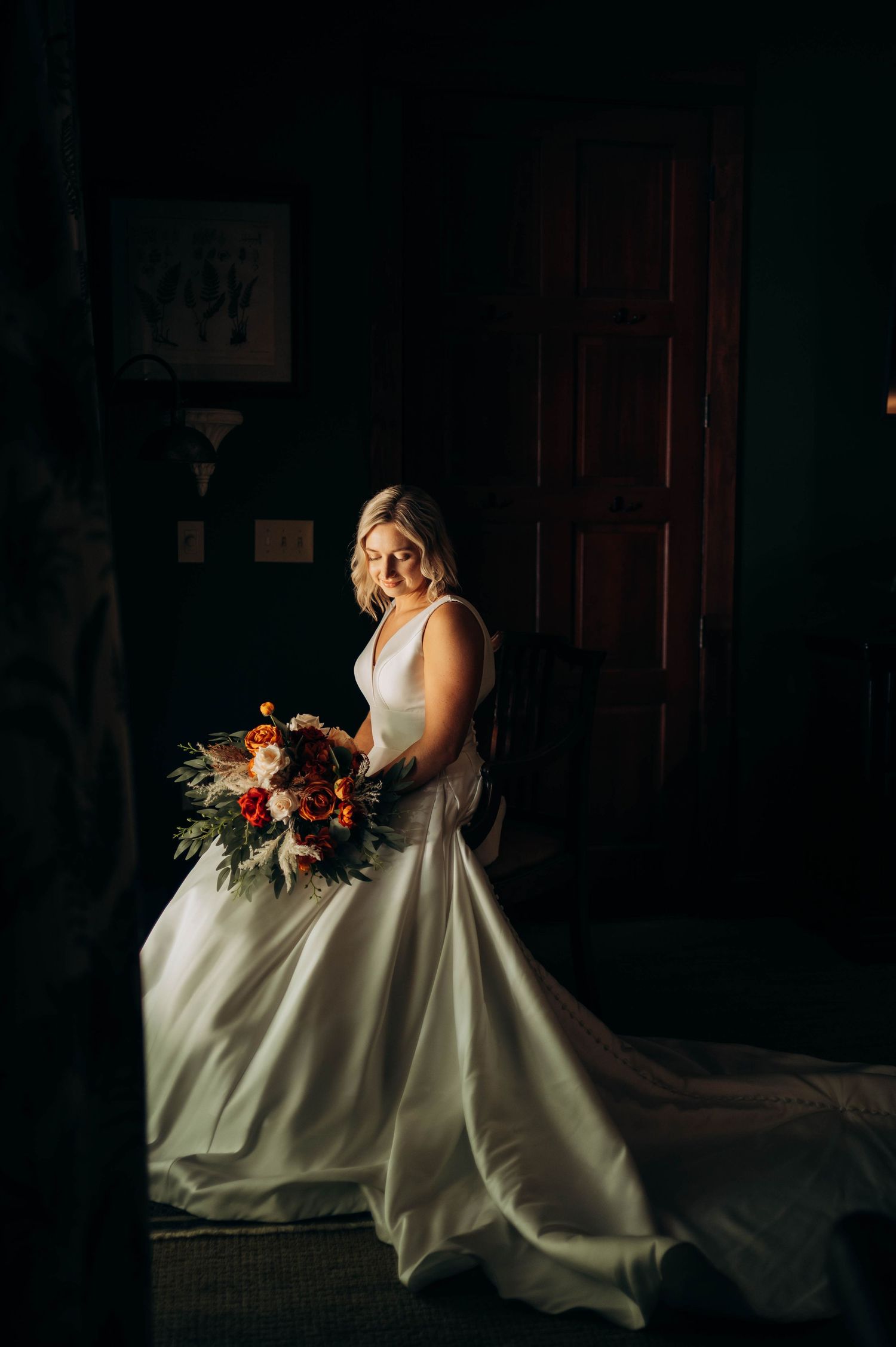 Bride in elegant white gown sits in dark room holding autumn bouquet.