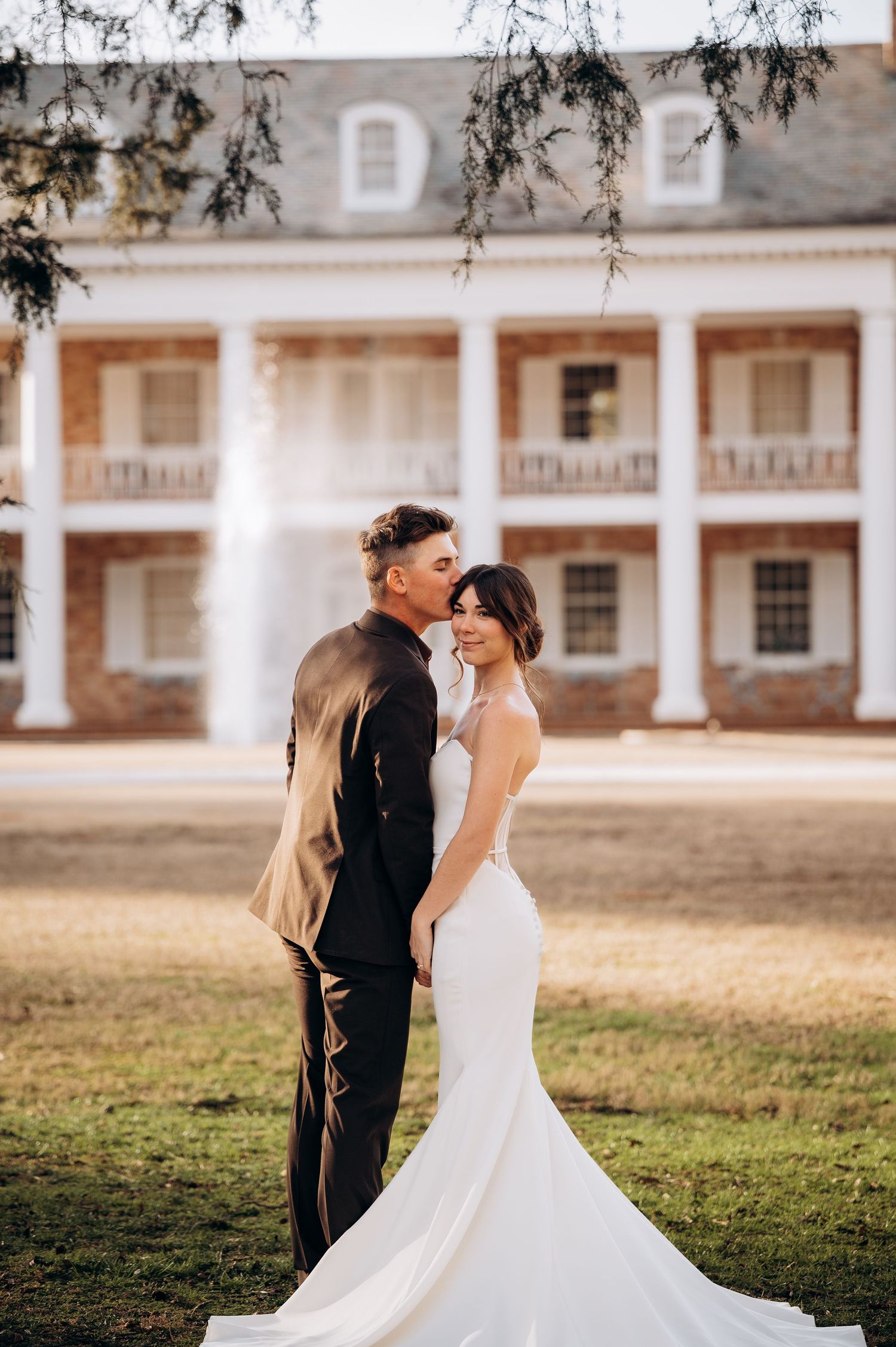 Romantic wedding couple embraces on a grassy lawn in front of a historic colonial-style building with white columns.