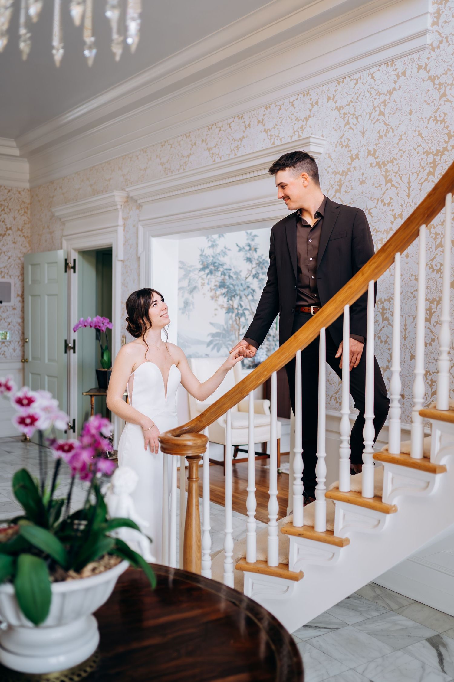 A romantic first look moment captured on an elegant staircase with white walls and a purple orchid in the foreground.