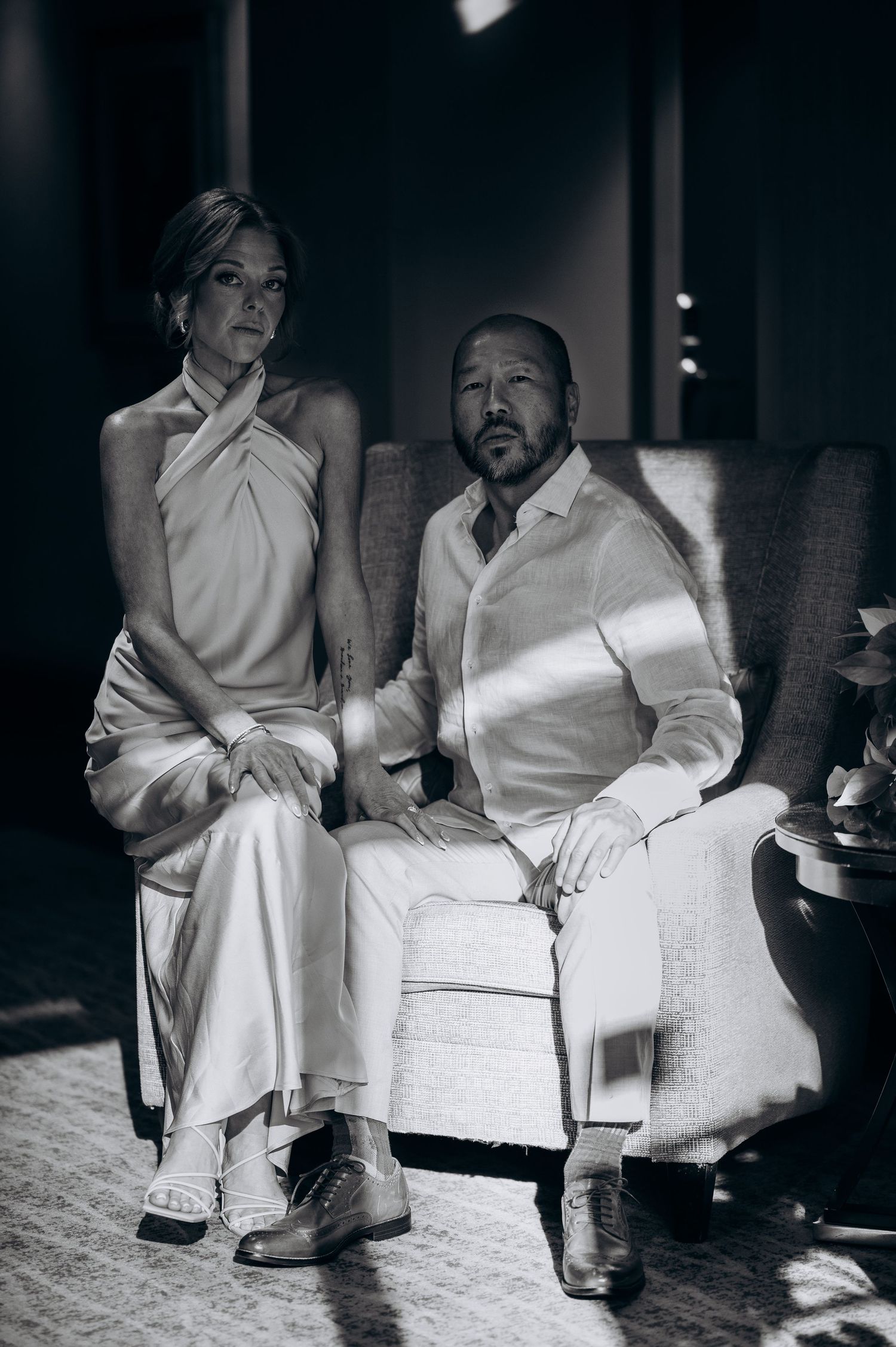 Elegant couple in white wedding attire seated together in a black and white portrait.