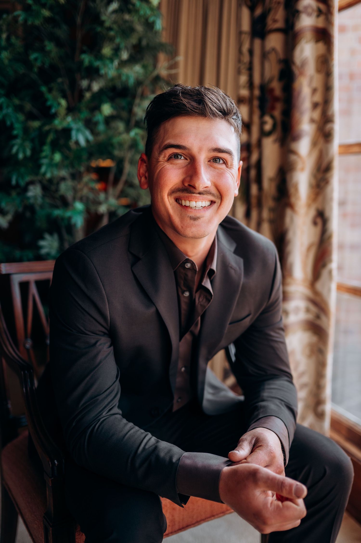Smiling groom in a dark suit and brown shirt seated near a window with greenery.