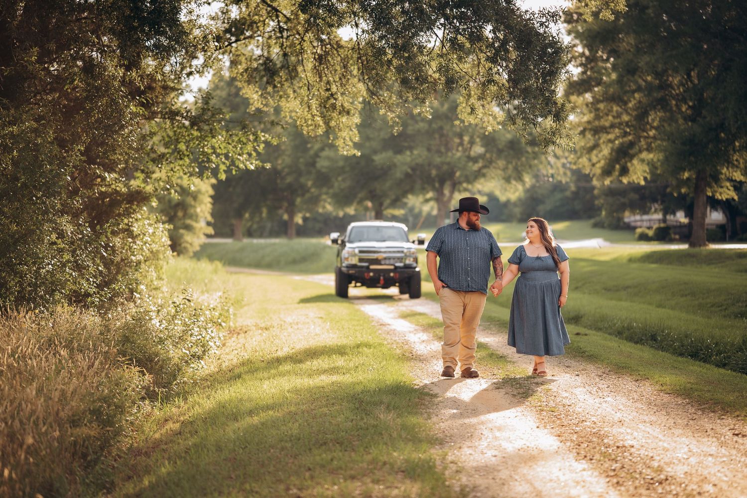 Couple walking hand in hand down a sunlit rural dirt path with a truck parked in the background.
