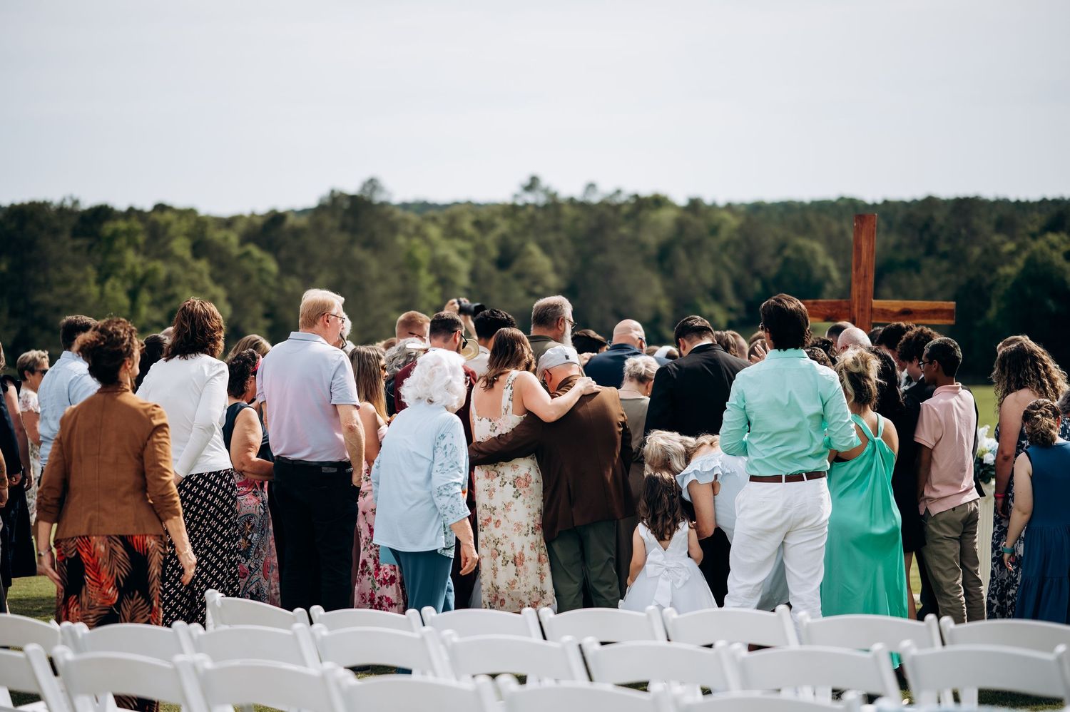 Wedding guests gathered at outdoor ceremony facing a wooden cross on a scenic green landscape.
