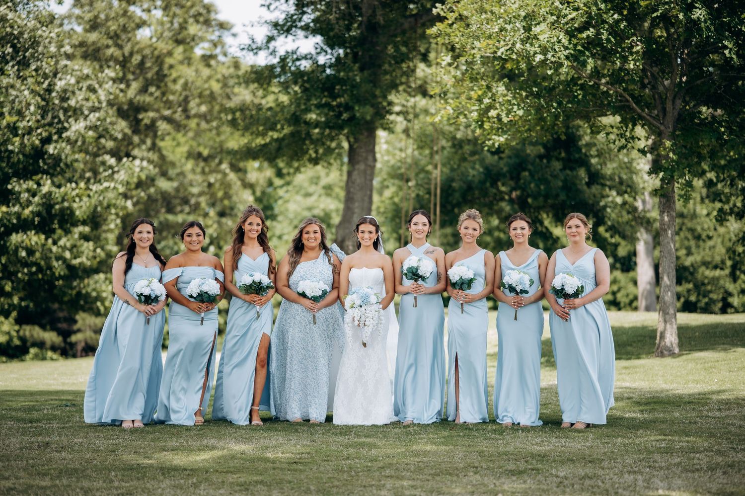 Bride in white gown poses with nine bridesmaids in light blue dresses on a green lawn surrounded by trees.