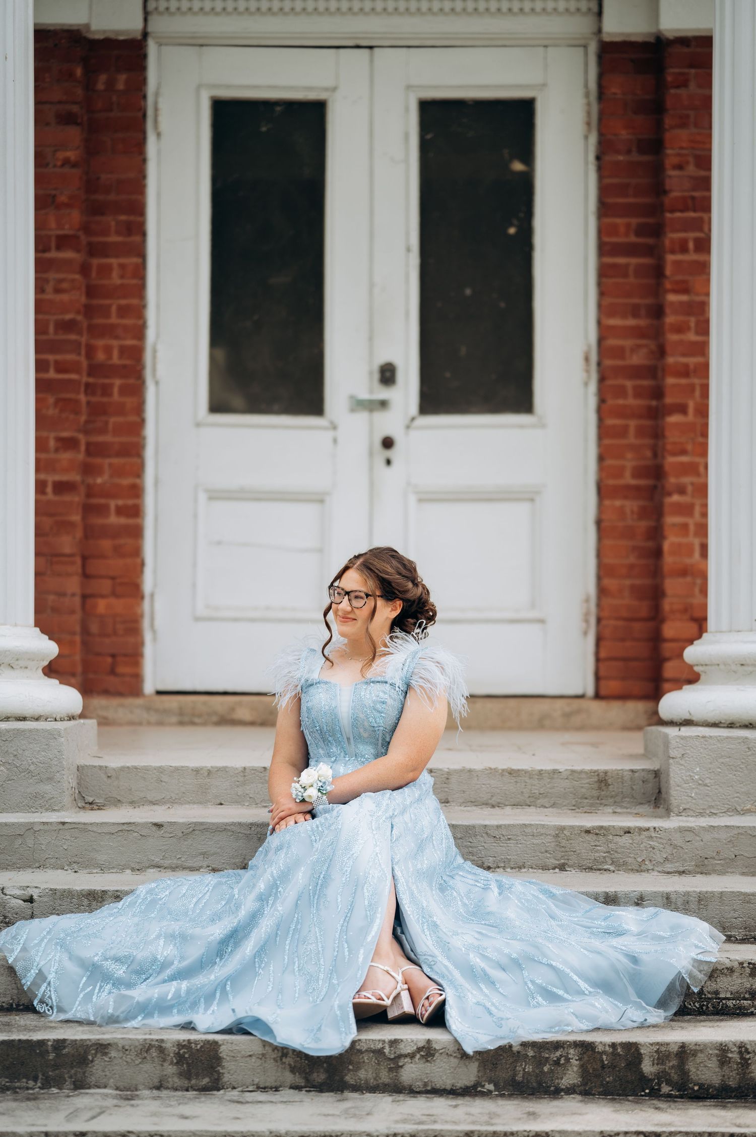 Young woman in light blue prom gown with wrist corsage sits on stone steps before white doors.