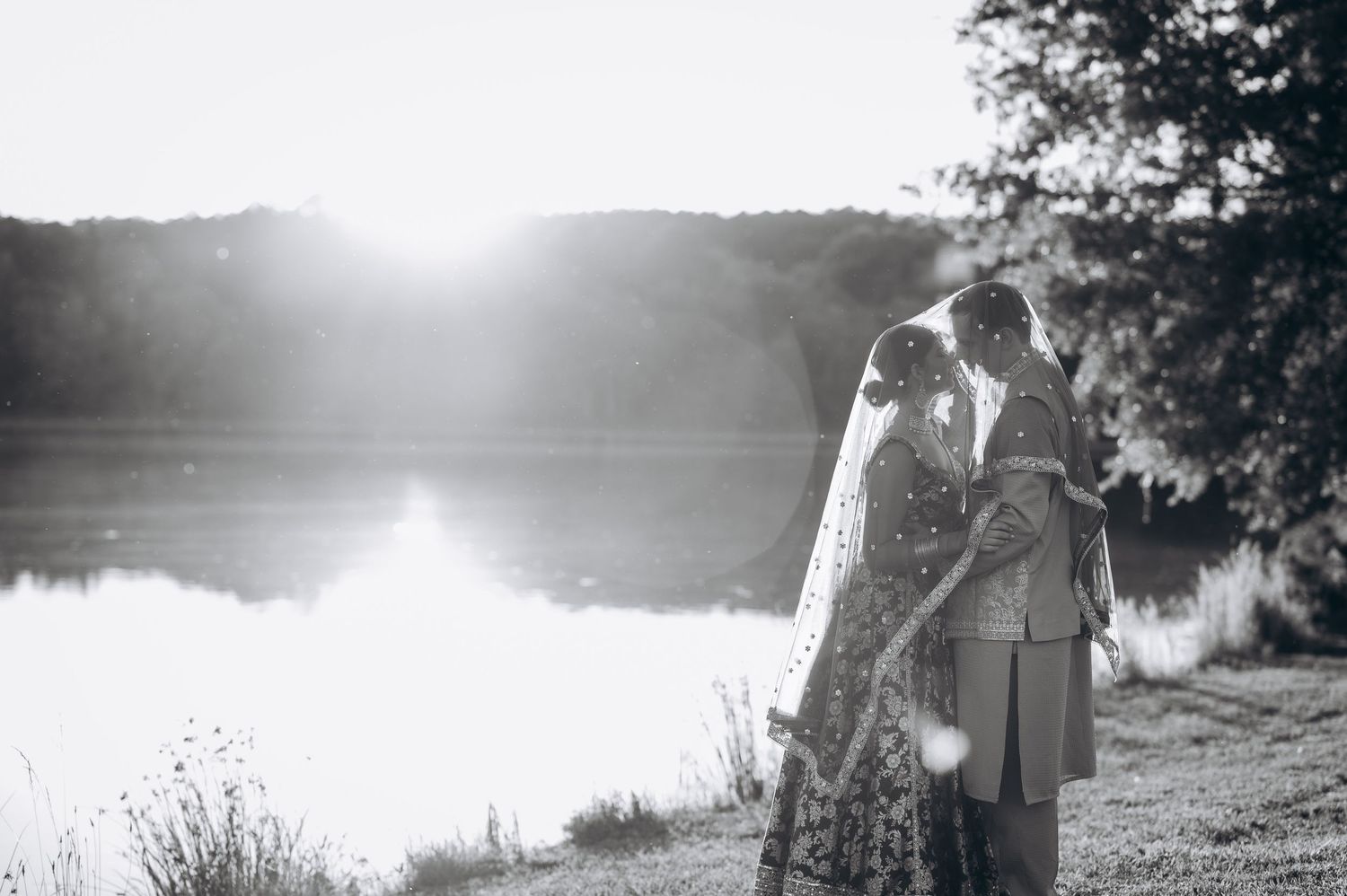 Black and white photo of a bride and groom sharing a romantic moment by a sunlit lake on their wedding day.