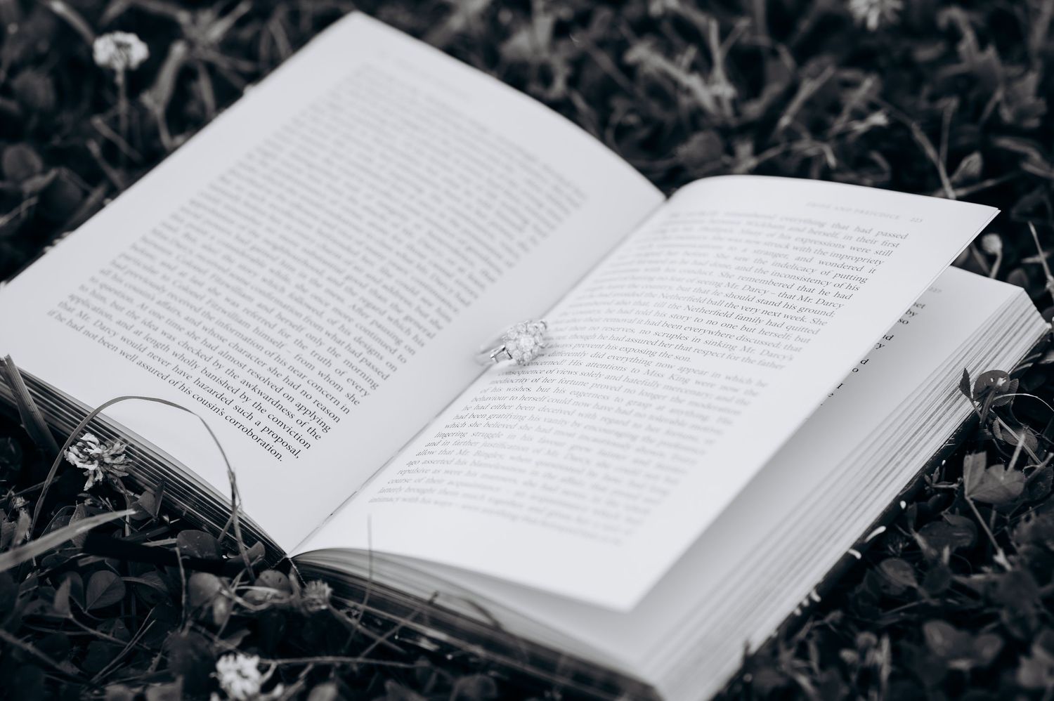 Open book with a diamond ring resting on its pages, lying on grass in black and white photography.