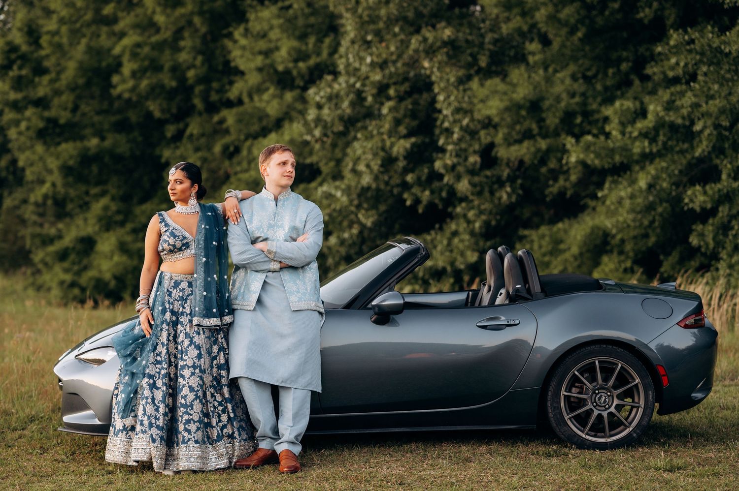 Couple in elegant Indian and Western attire posing beside a gray Mazda MX-5 Miata convertible outdoors.