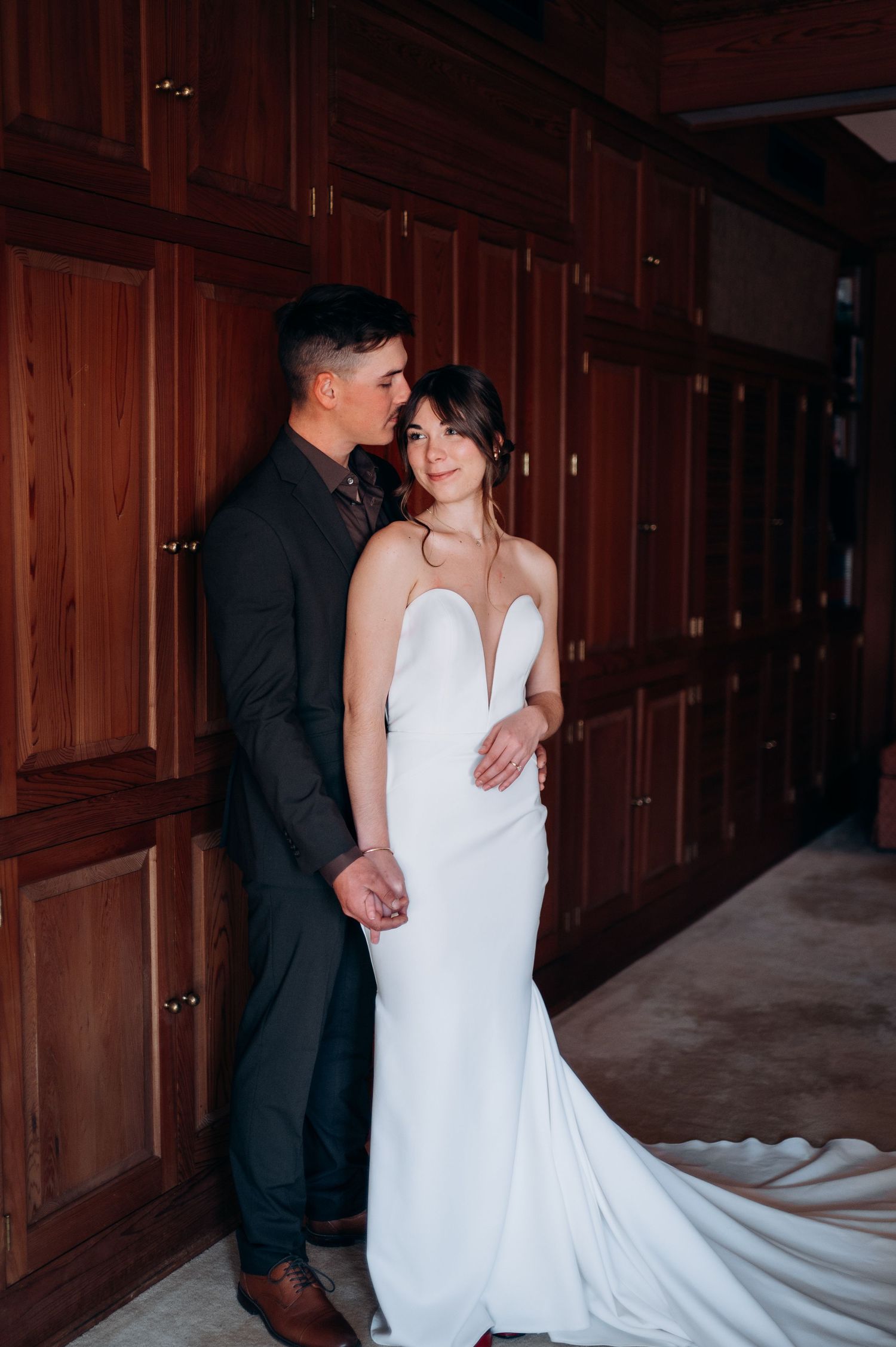 Couple in wedding attire embracing against rich wood-paneled wall.