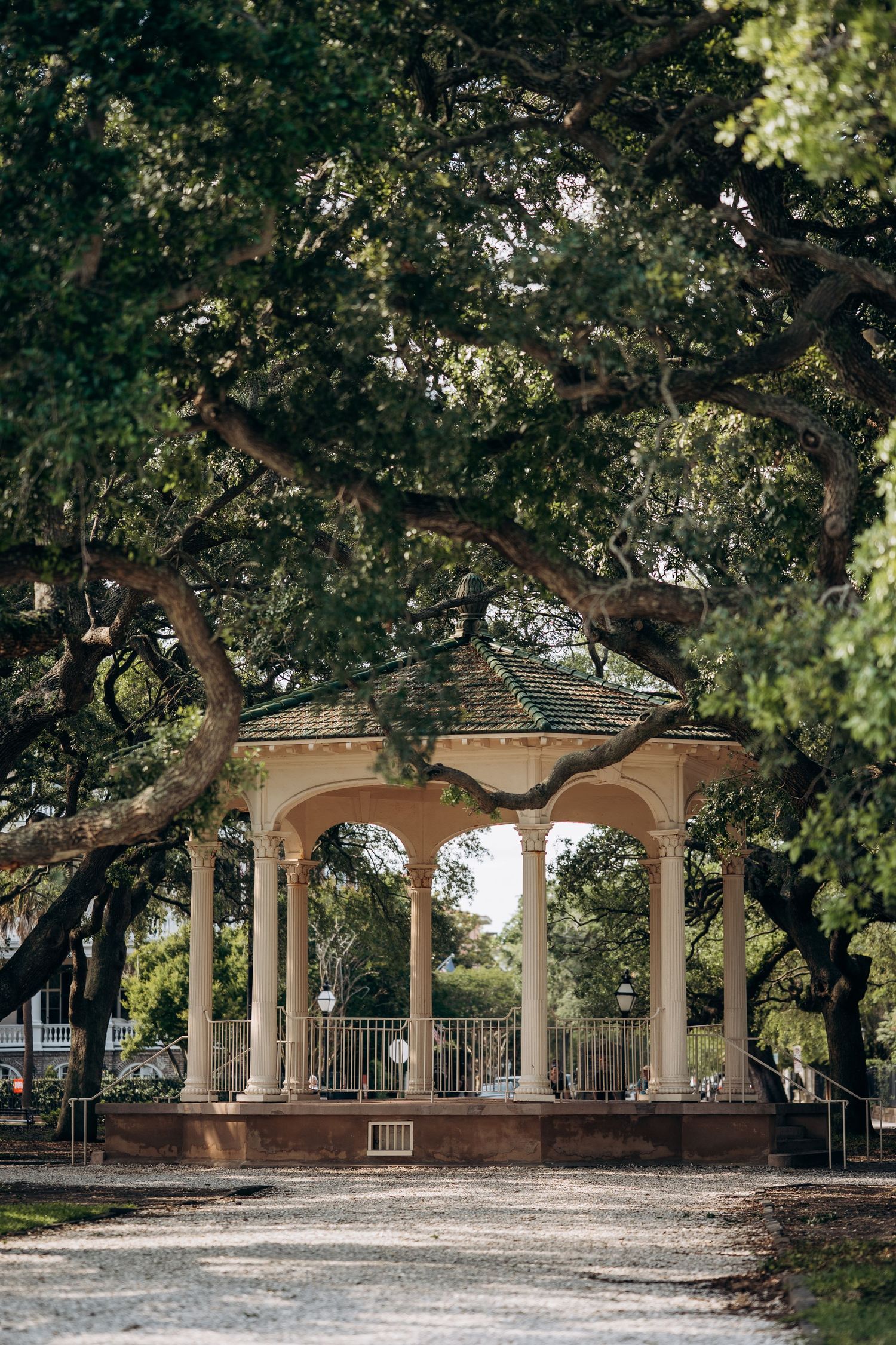 A elegant white gazebo with arched columns nestled beneath sprawling oak trees in a lush park.