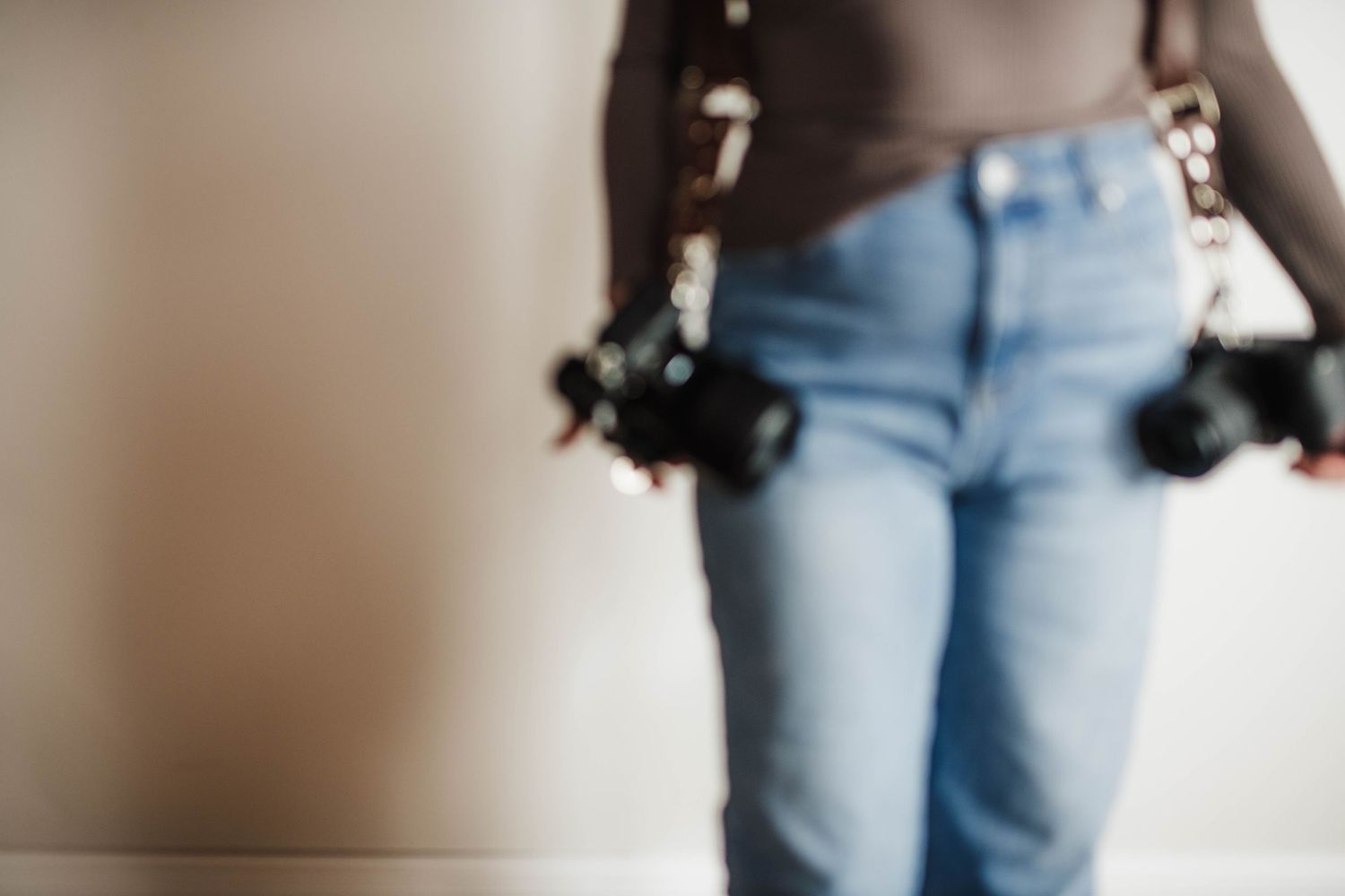 Photographer in blue jeans and black top holds camera while standing against white wall.