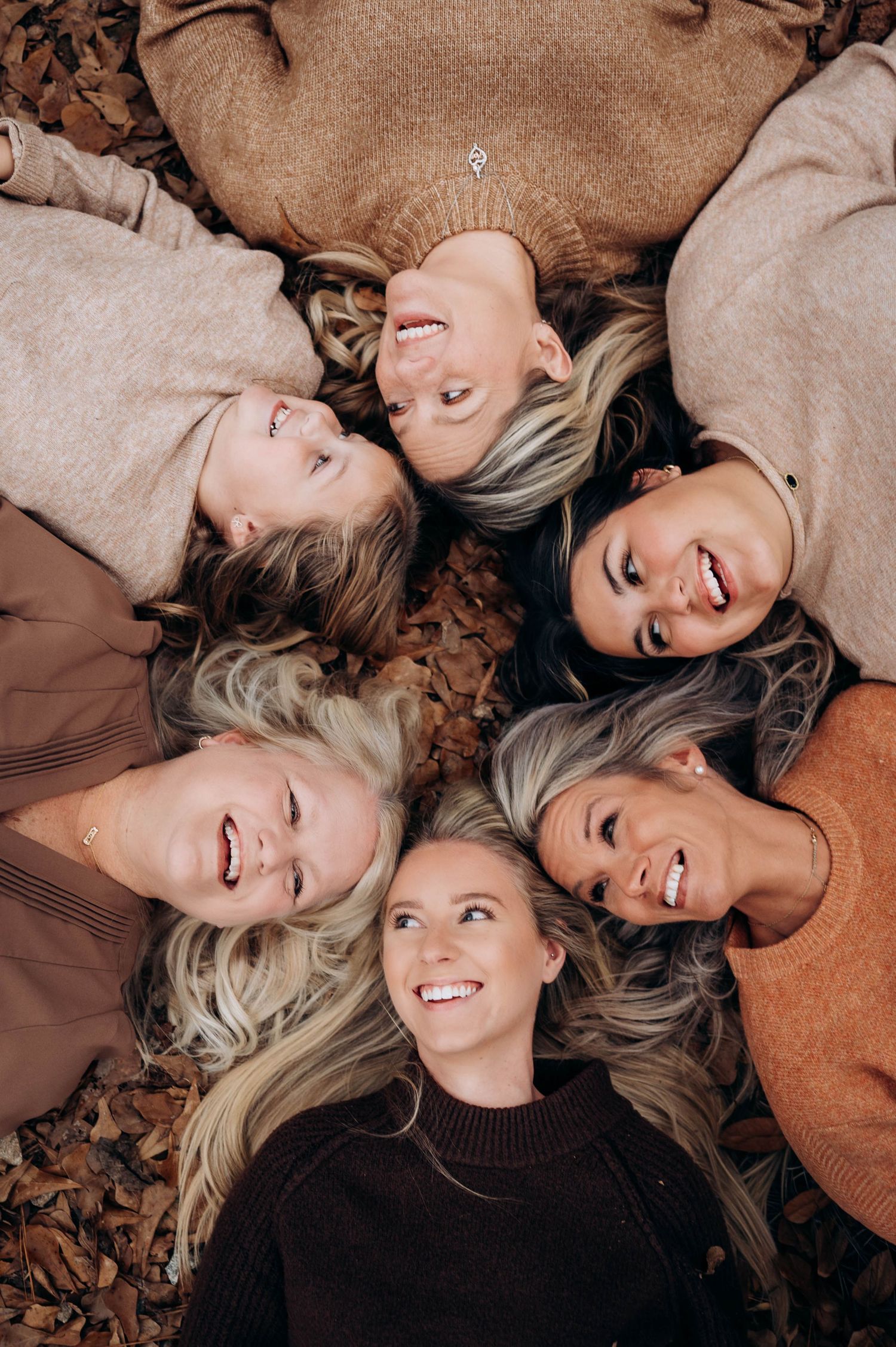Friends lying in circle on autumn leaves wearing cozy neutral sweaters.