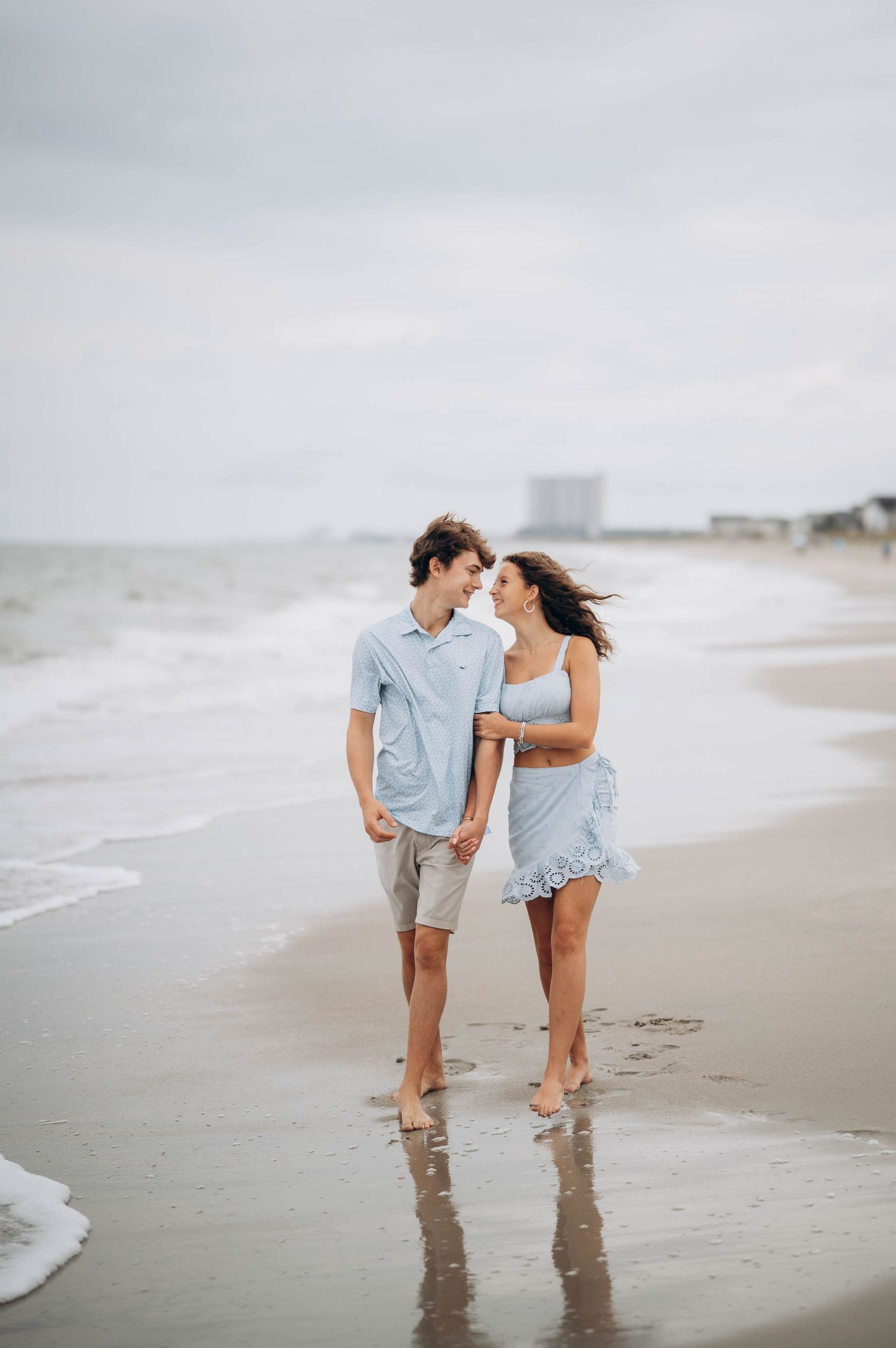 A couple walking barefoot together along a misty beach shoreline on an overcast day.