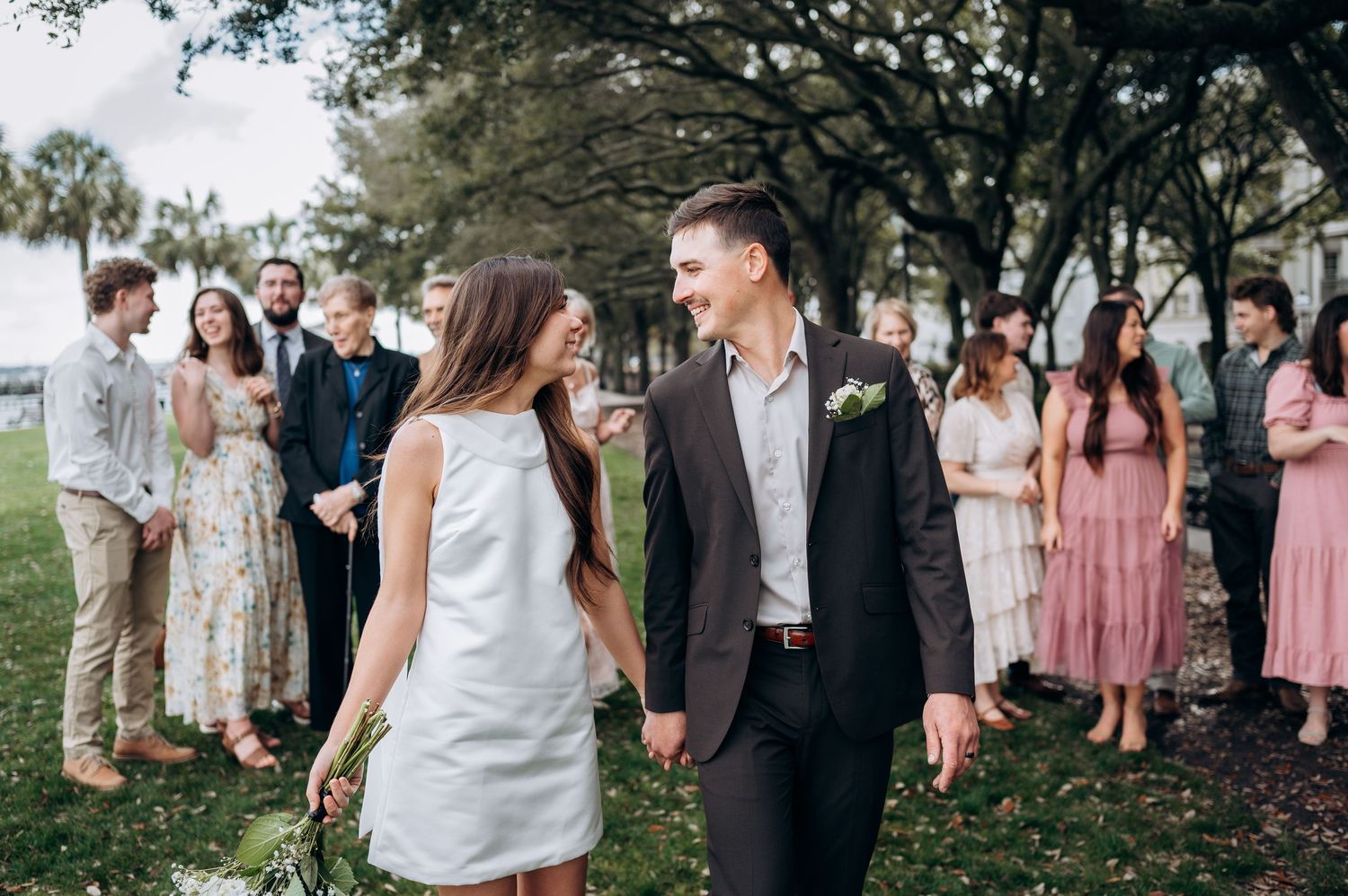 Wedding party members gather outdoors under trees wearing pink dresses and formal attire.