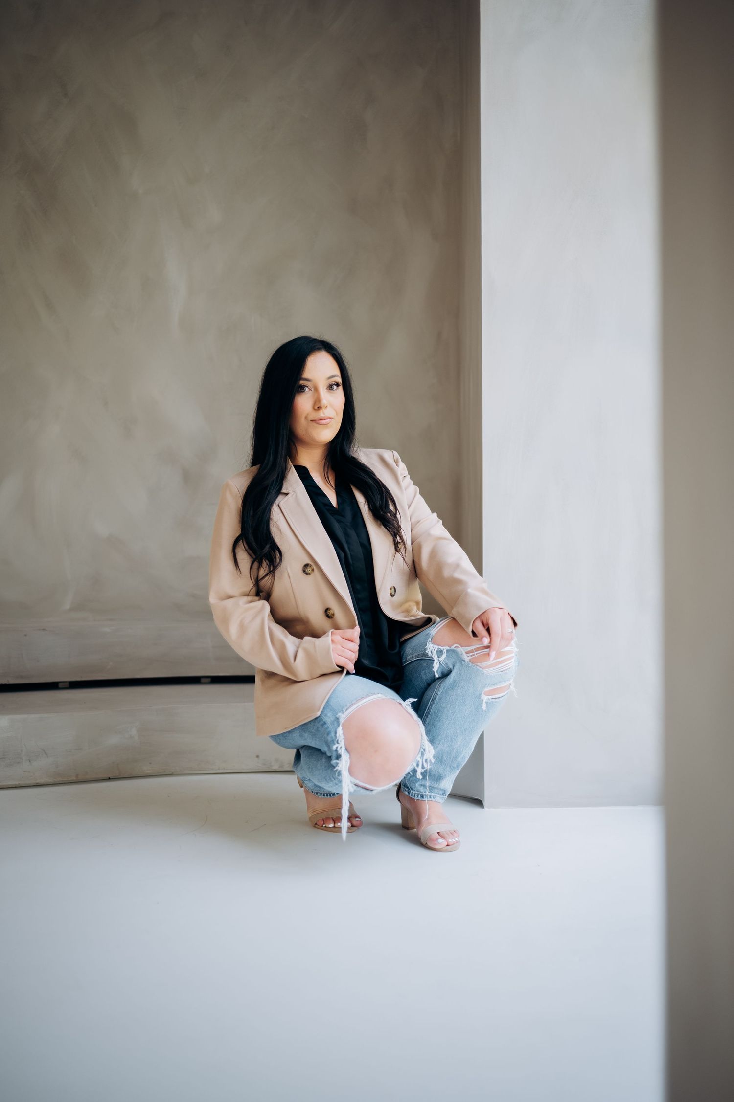 A person in a beige blazer and ripped jeans poses crouching against a minimalist gray wall.