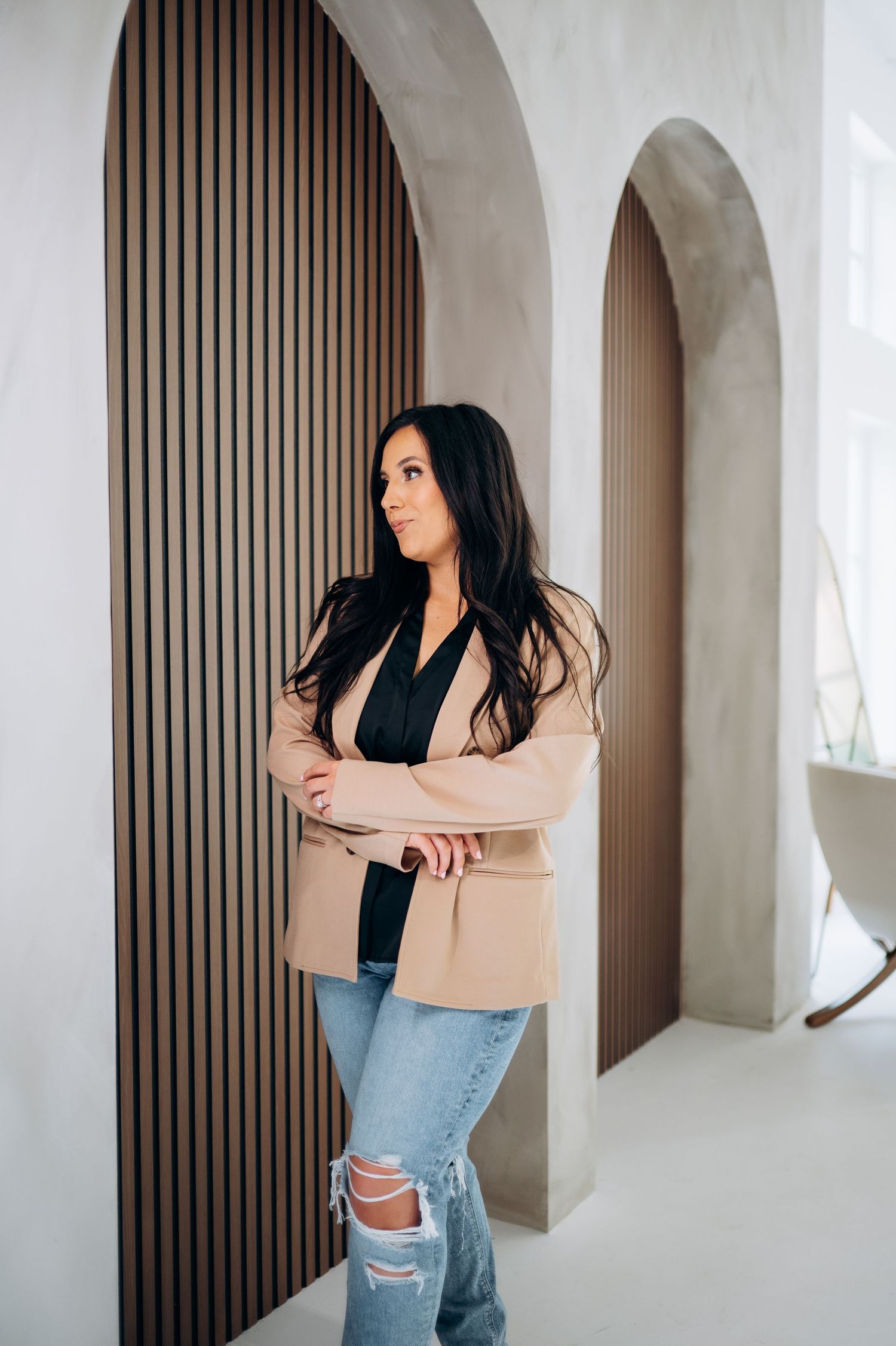 Woman in beige blazer and ripped jeans poses against white wall with arched doorways and wood paneling.