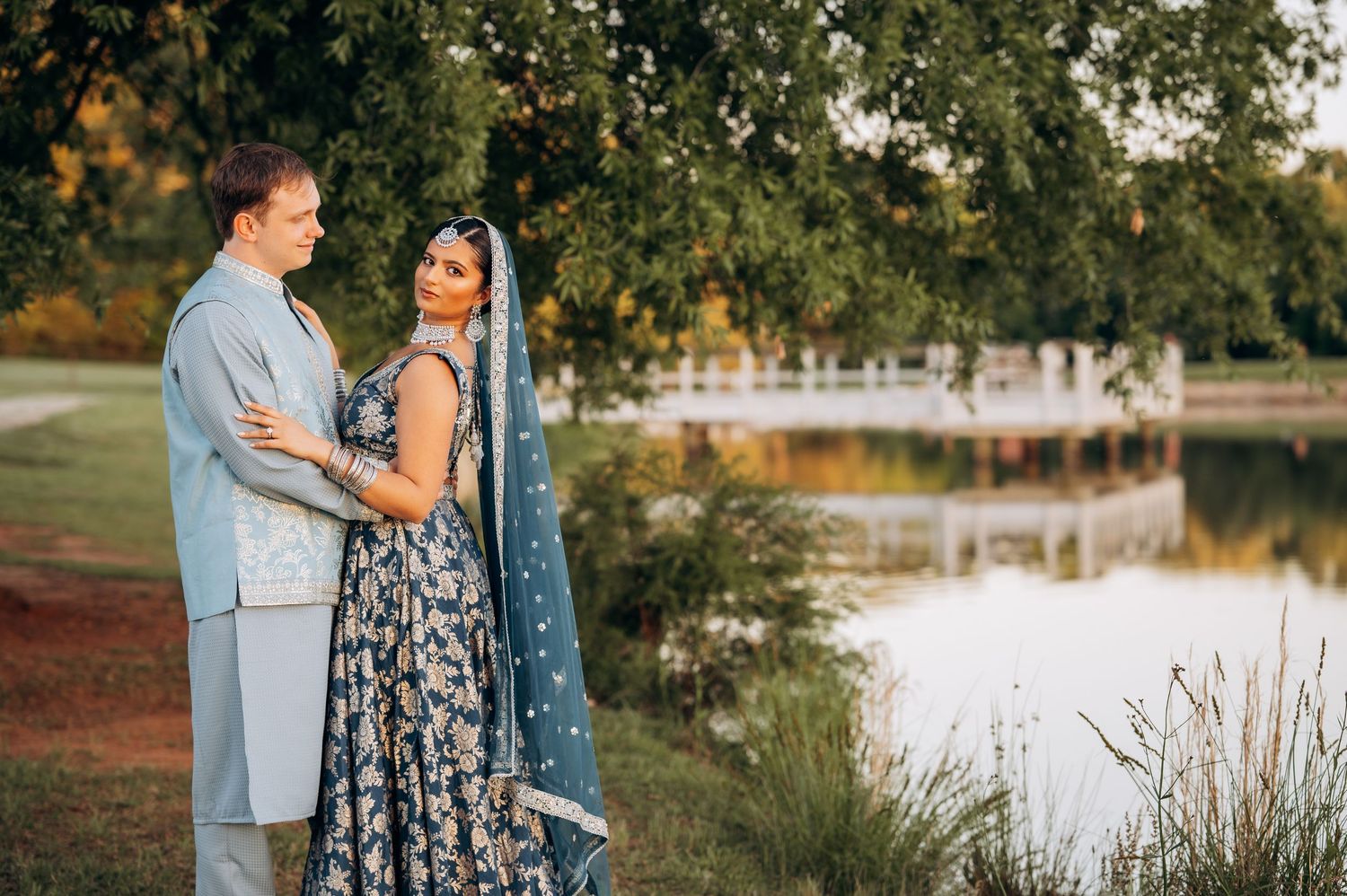 Couple in elegant Indian wedding attire poses by a serene lake surrounded by lush green trees at golden hour.