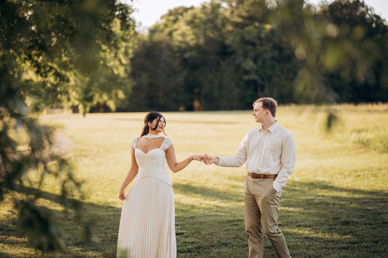 Couple holding hands in a sunlit green field, bride in white dress and groom in khaki, golden hour wedding photo.
