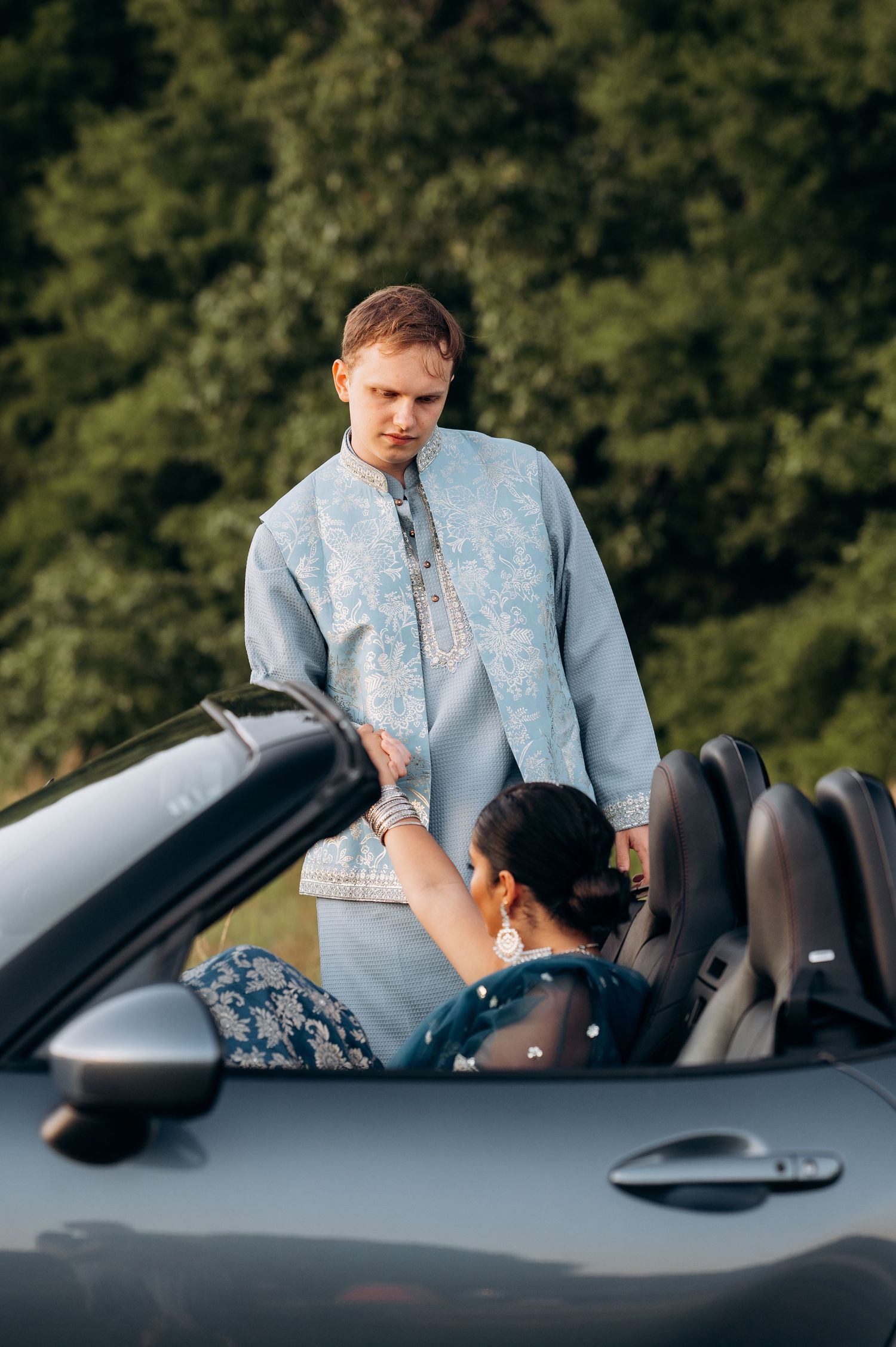Man in blue kurta holds hands with woman in convertible car, both dressed in elegant Indian attire outdoors.