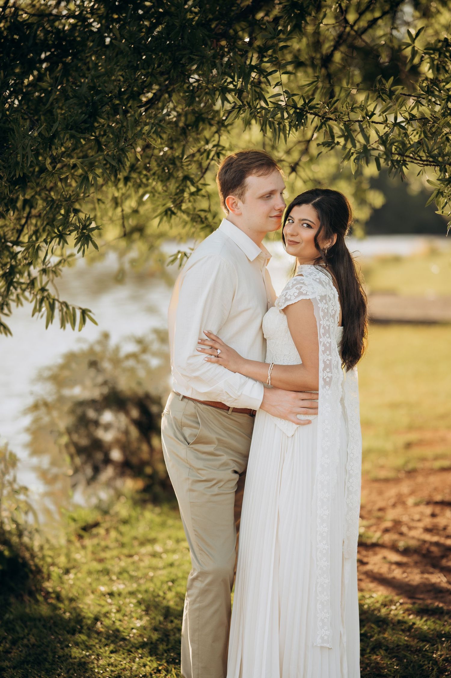 Couple in white wedding attire embracing under willow tree by a sunlit lake, romantic outdoor ceremony photo.