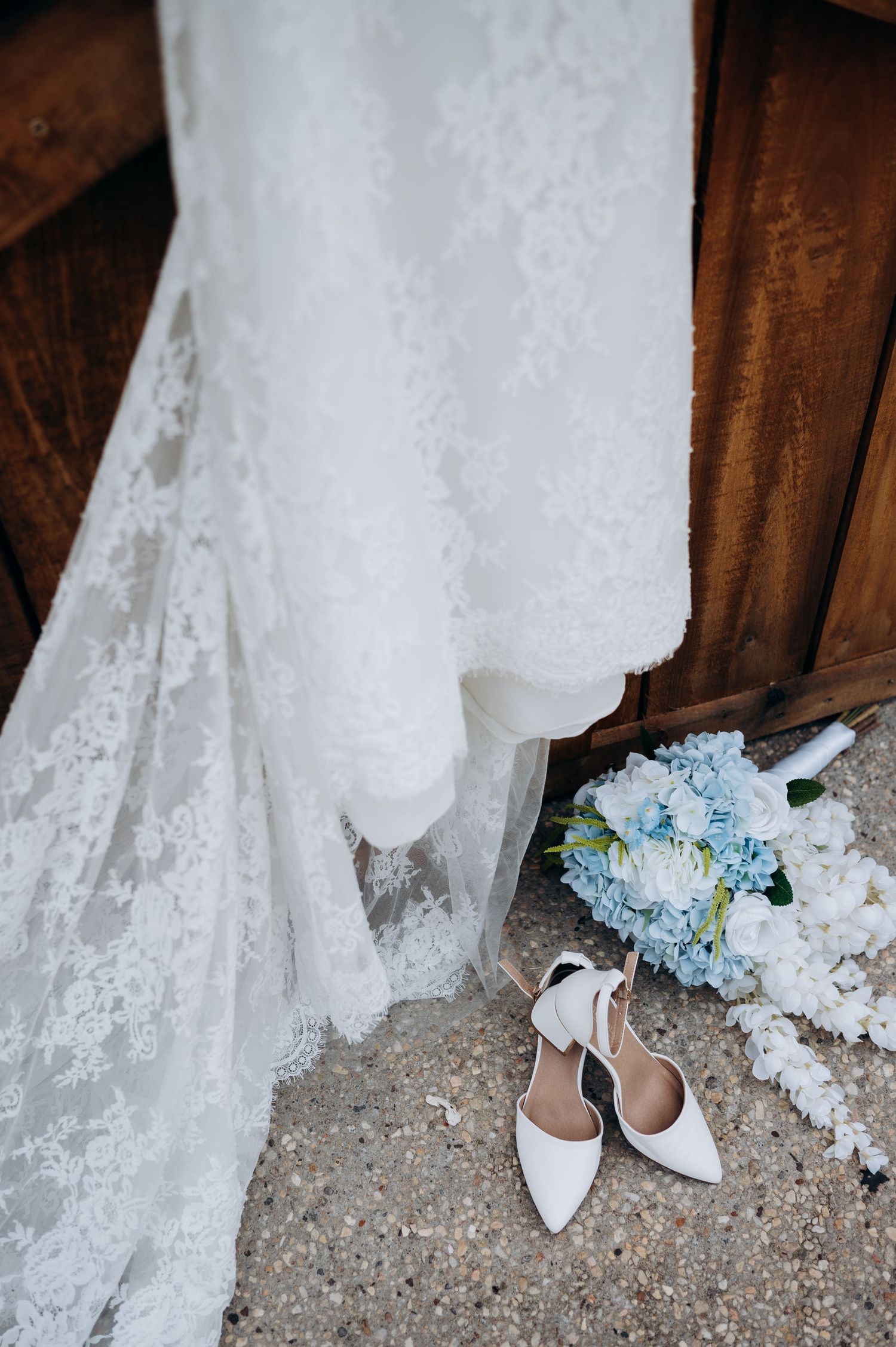 White lace wedding dress hanging beside bridal bouquet of blue hydrangeas and white roses with elegant pointed-toe heels.