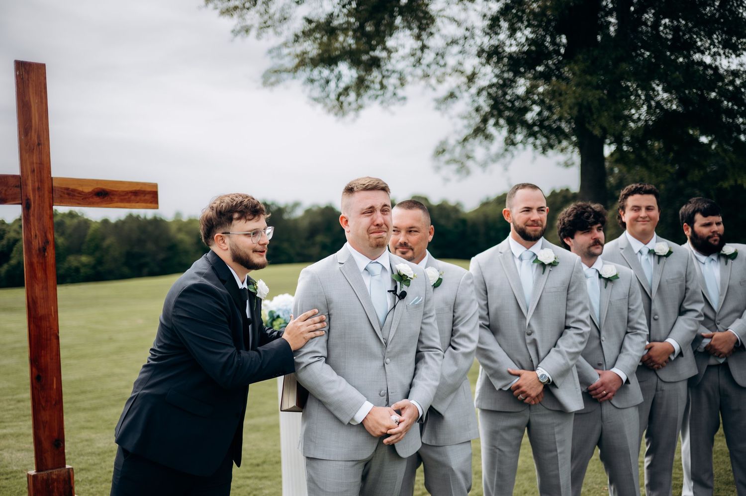 Groom in grey suit shares emotional moment with officiant beside wooden cross at outdoor ceremony.