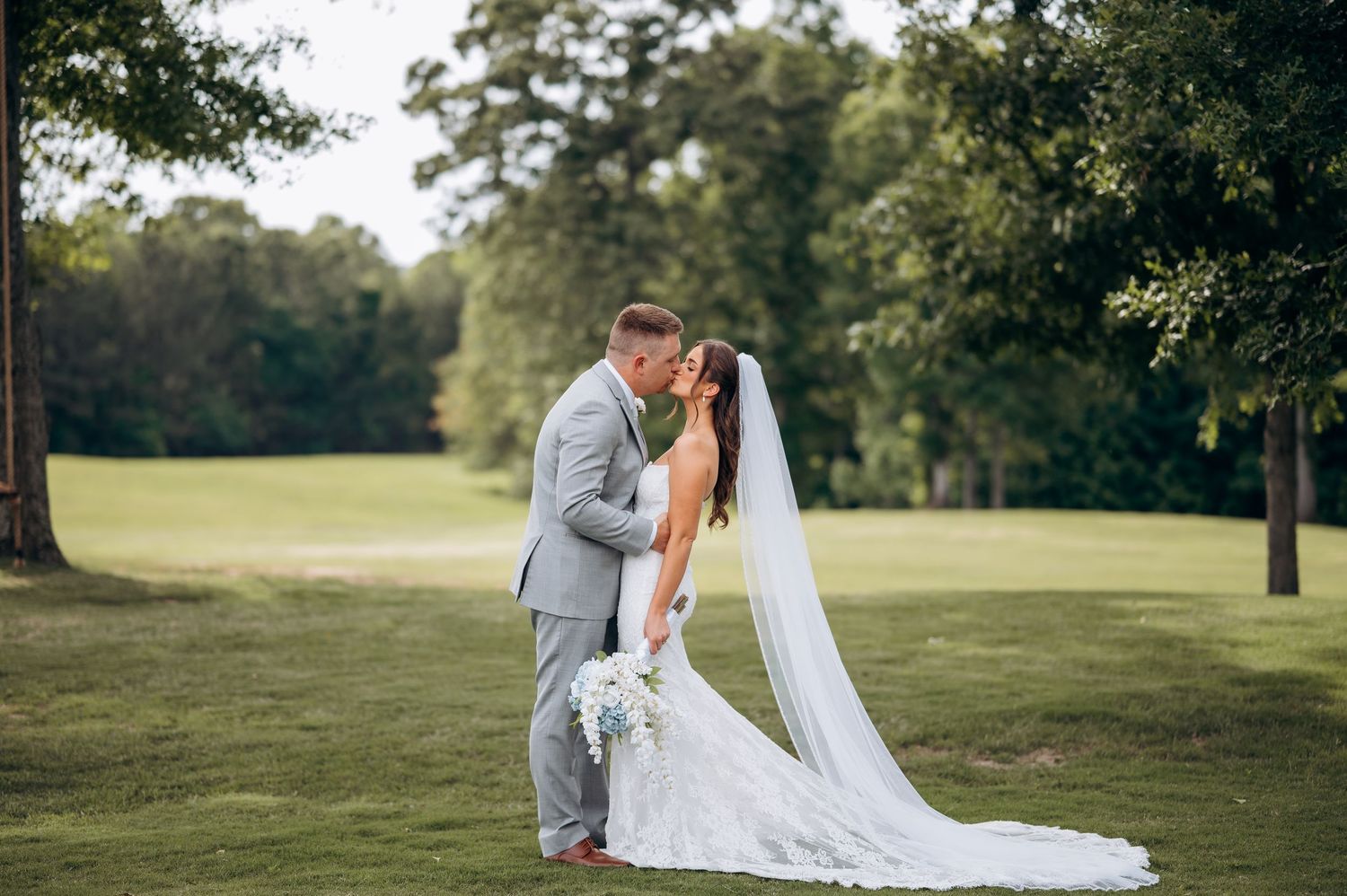 Bride and groom sharing a romantic kiss on a lush green golf course, bride in lace gown with cathedral veil.